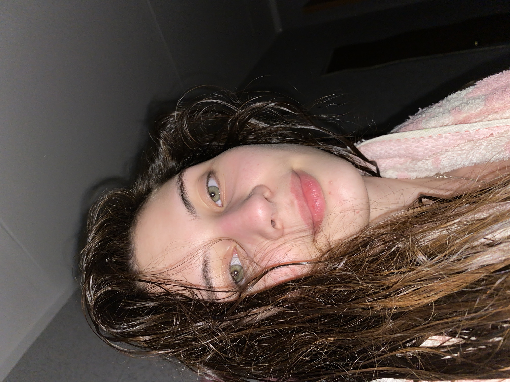 A young woman lies on her back. Her hair is wet and dark brown. Her eyes are hazel. She has a fair complexion and a slight smile. She is wearing a pink and white patterned robe or towel. The background consists of a grey wall and ceiling, and a dark area in the upper right corner. There is no indication of the city of Votkinsk, Russia.