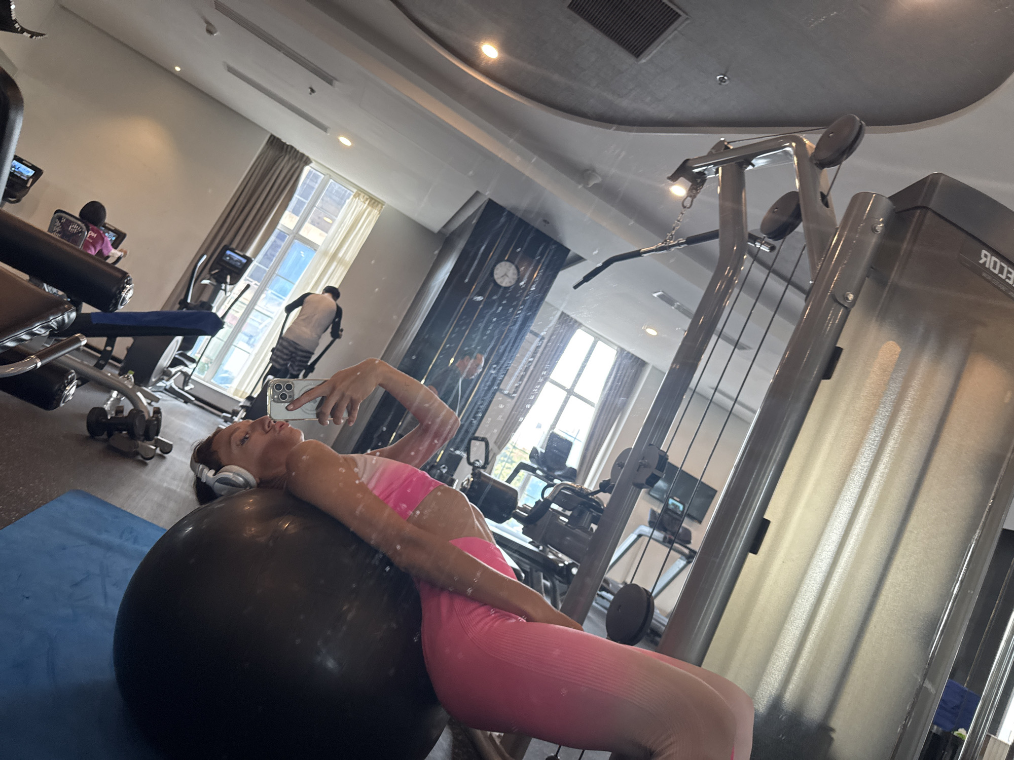 The image appears to be taken in a gym located in Đà Nẵng, Vietnam. A woman wearing pink athletic wear and white headphones is lying supine on a large black exercise ball, holding a smartphone up to take a photo of herself. Gym equipment, including weight machines and treadmills, are visible in the background. People can be seen exercising on some of the machines. The room has large windows with light-colored curtains.