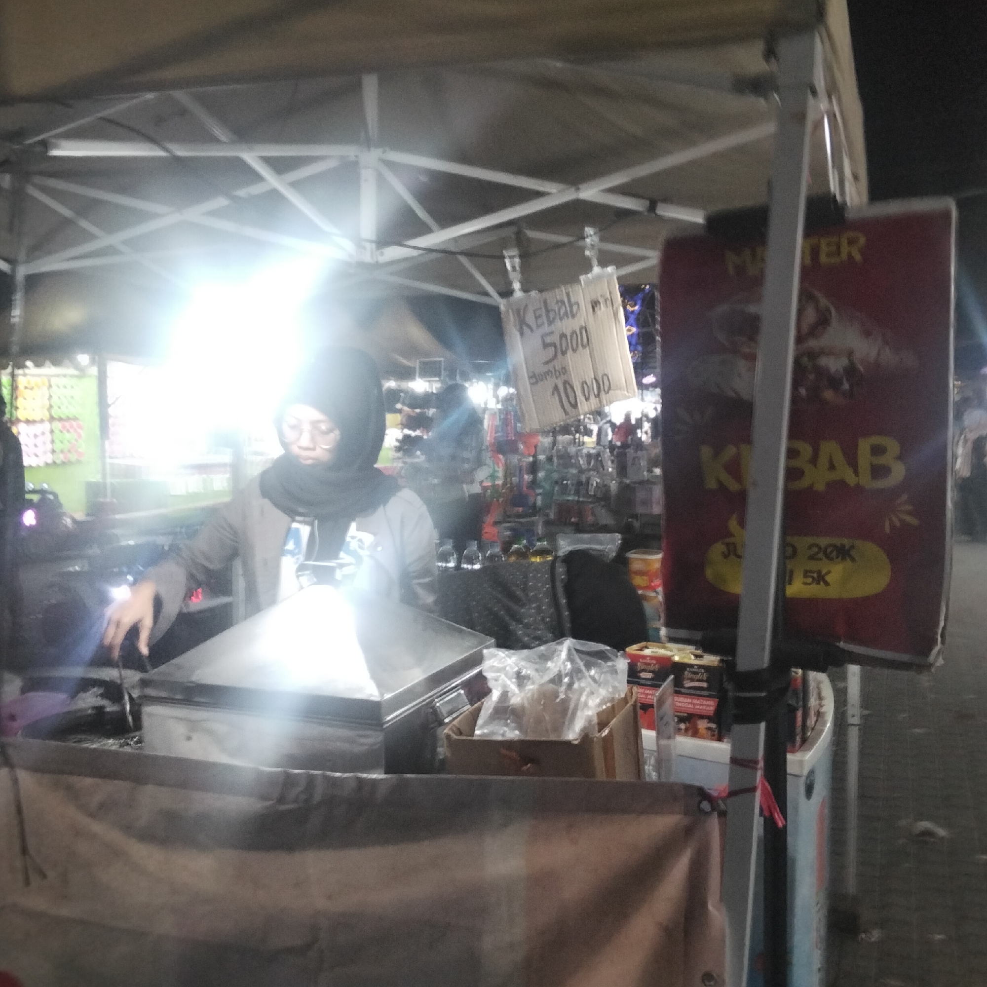 This is a medium-shot, eye-level, night-time photograph of a street food stall in Cisaat, Indonesia. A woman wearing a black hijab and glasses, and a grey jacket, is tending to the stall. She is leaning over a metal food warmer and appears to be working with tongs. The stall is covered by a white canopy, illuminated by bright overhead lights, creating a lens flare effect in the upper left portion of the image.
In the background, other stalls and people are visible, suggesting a busy night market or food court setting. A sign made of cardboard is hanging to the right of the woman, with handwritten text and prices for "Kebab". Another larger, red sign with yellow lettering is also visible to the right, with the word "Kebab" prominently displayed, along with prices like "20K" and "5K". Various bottles and containers are on display on shelves behind the woman. The overall scene depicts a typical night-time food vendor operation.