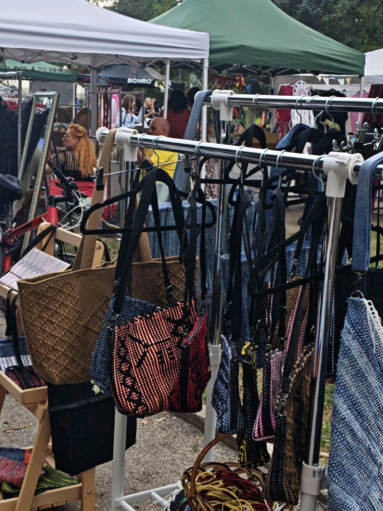 An outdoor market stall is depicted, featuring racks of handbags and various textile items. In the foreground, a large woven tote bag in a golden-brown hue is prominently displayed. To its right, a smaller bag with a black and orange patterned weave hangs. Behind these, a rack holds numerous handbags with black straps, showcasing a variety of colors and textures, including blue, black, and patterned fabrics. A large, textured blue and white woven textile is visible on the far right. In the background, people are seen browsing under tents, with the green canopy of a tent visible overhead. A white tent structure is also present. The ground appears to be paved with small stones or gravel. A bicycle with a red frame is partially visible on the left. The overall scene suggests a vibrant market environment. The location is identified as Vinnytsia, Ukraine.