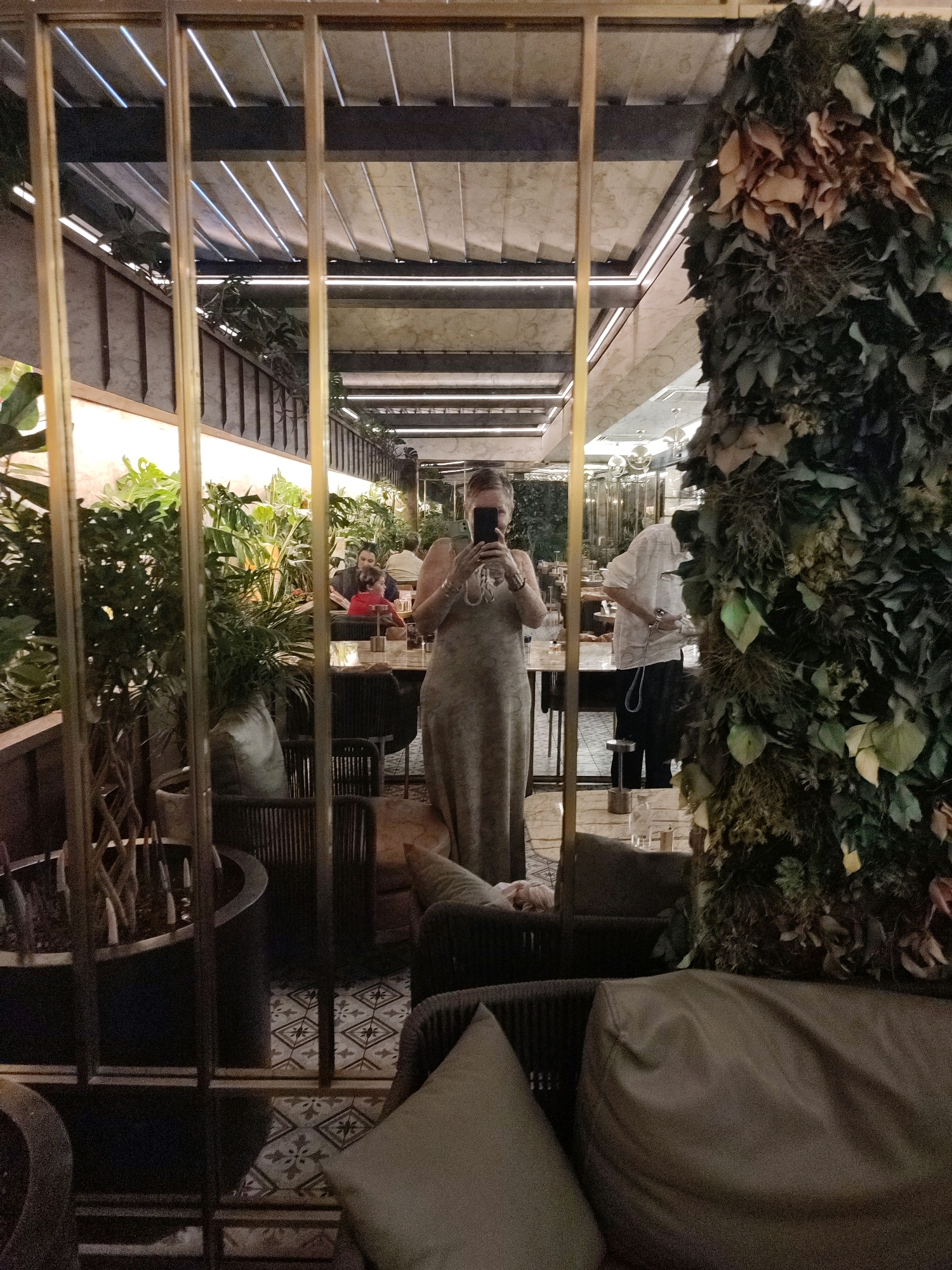A woman is taking a selfie in a mirror in what appears to be a restaurant or cafe in Istanbul, Turkey. She is wearing a light-colored, long dress and is holding a smartphone. The reflection shows other patrons seated at tables and a living green wall feature. The interior design includes modern furniture, plants, and decorative lighting.