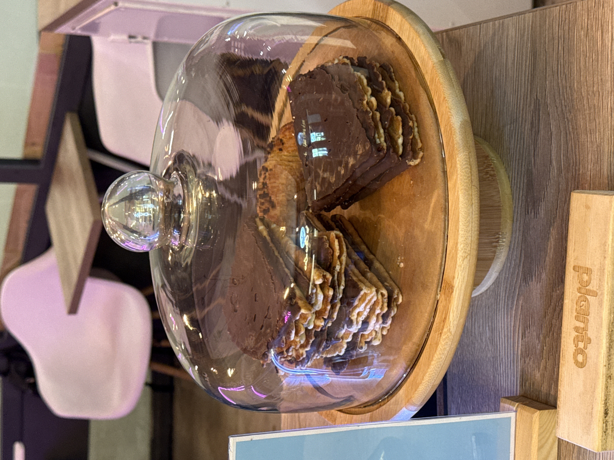 The image depicts a close-up view of a glass dome covering a wooden cake stand, which holds a stack of chocolate-covered wafers and a croissant. The background shows a cafe setting with pink chairs and wooden tables. The lighting suggests an indoor environment, possibly a bakery or cafe. There are no people visible in the image. A wooden sign with the embossed text "Planto" is partially visible in the foreground. Based on the context of "Planto" likely being a cafe or bakery name, this scene could be located in Moscow, Russia.