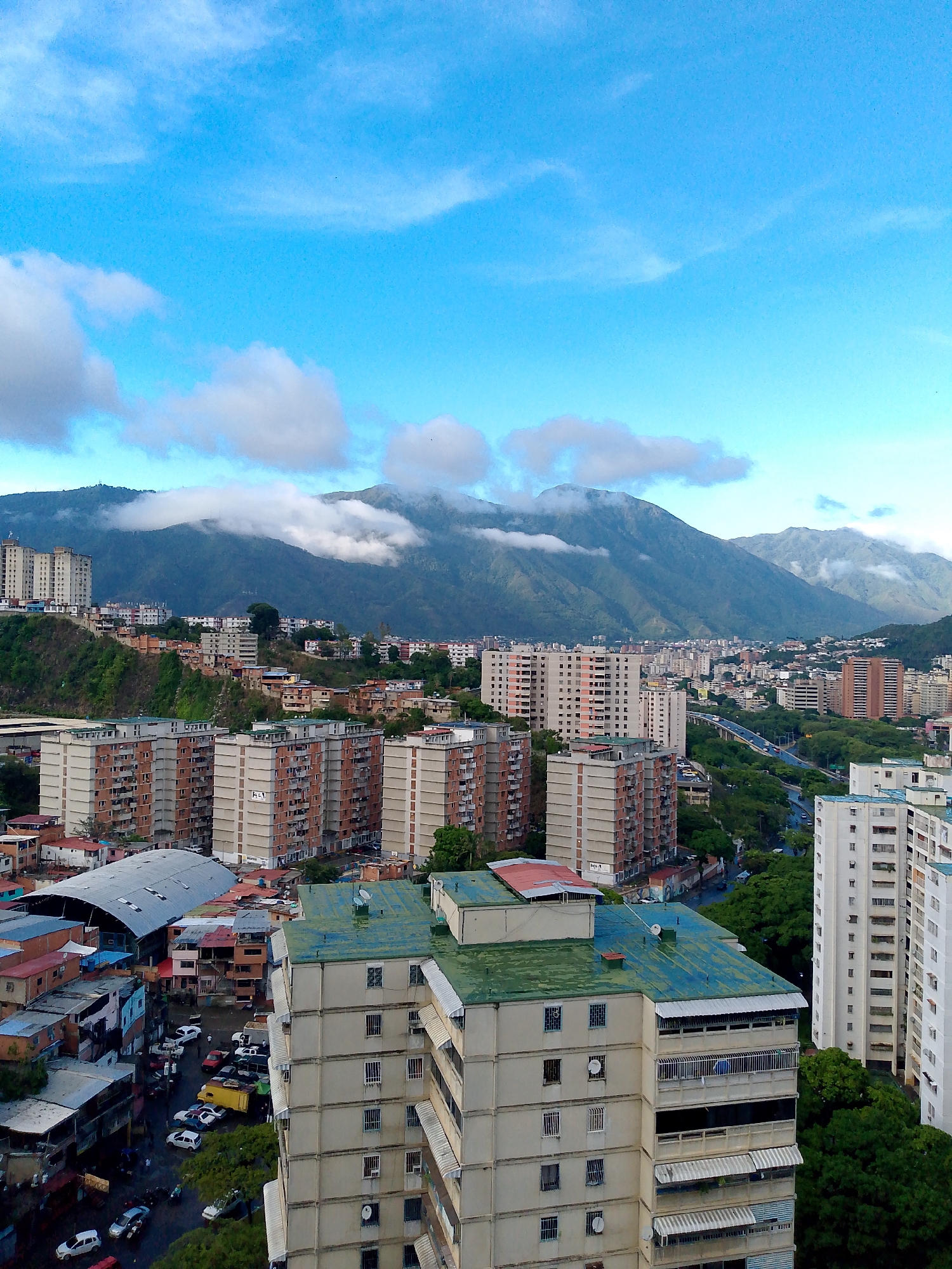This is an aerial view of a densely populated urban area in Caracas, Venezuela, with a mountain range in the background. The foreground features several multi-story apartment buildings, some with light-colored facades and others with reddish-brown brickwork. Many of these buildings have balconies with railings and awnings.
Further into the city, there are more apartment buildings of varying heights and architectural styles, interspersed with trees. A highway overpass with cars driving on it is visible, suggesting a bustling transportation network. The mountains in the background are partially obscured by clouds, with some areas appearing green and others rocky.
In the lower left of the image, there's a collection of smaller, older buildings with colorful roofs and walls, and what appears to be an outdoor market or covered area with a large, metallic roof structure. Cars are parked in an open area, and some people are visible walking around. The overall impression is of a vibrant, active city nestled against a dramatic natural landscape.