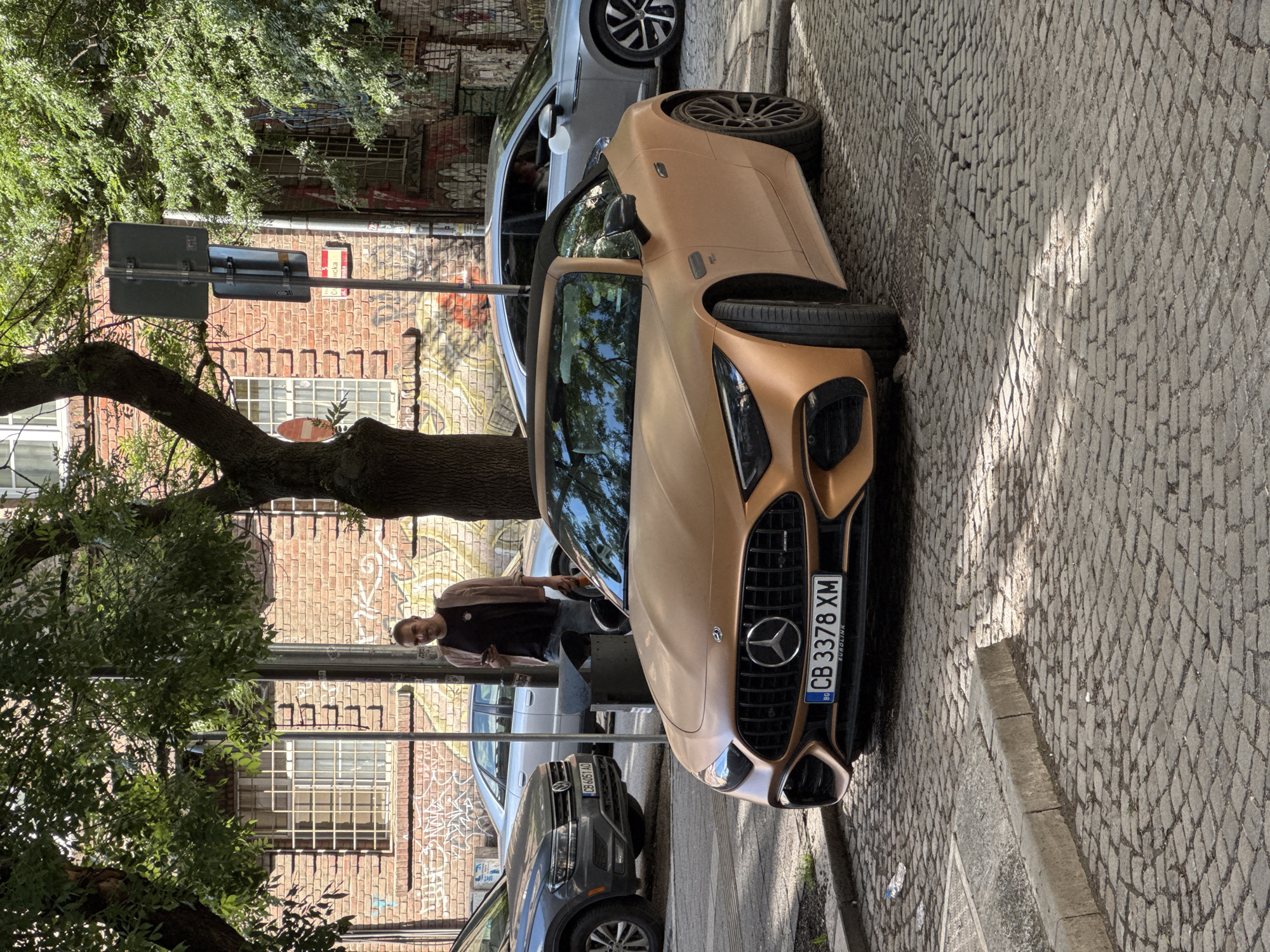 A photograph of an urban scene in Sofia, Bulgaria, depicts a matte gold-colored Mercedes-AMG GT parked on a cobblestone street. The license plate reads "CB 3378 XM" and is clearly visible. To the left of the Mercedes, a man stands partially obscured by a tree and a street sign. He is dressed in a black shirt and a light-colored jacket. Behind the Mercedes, a silver car is parked, and behind that, a gray Volkswagen Tiguan. The background consists of a brick building with graffiti and barred windows. The scene appears to be taken during daylight hours with shadows cast onto the cobblestone street.