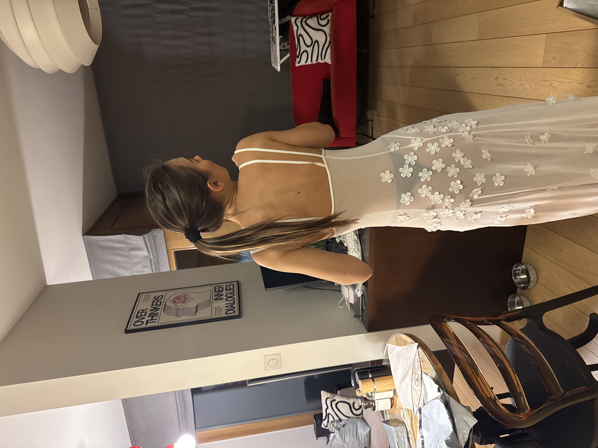 A woman stands indoors wearing a white dress with floral appliqués. Her hair is pulled back in a ponytail. The location appears to be an apartment in Warsaw, Poland.
The apartment features wooden floors, a red chair with a patterned pillow, and a poster with the words "Over Thinkers Inner Dialogues" on the wall. A modern light fixture hangs from the ceiling. A wooden chair is also visible in the foreground.