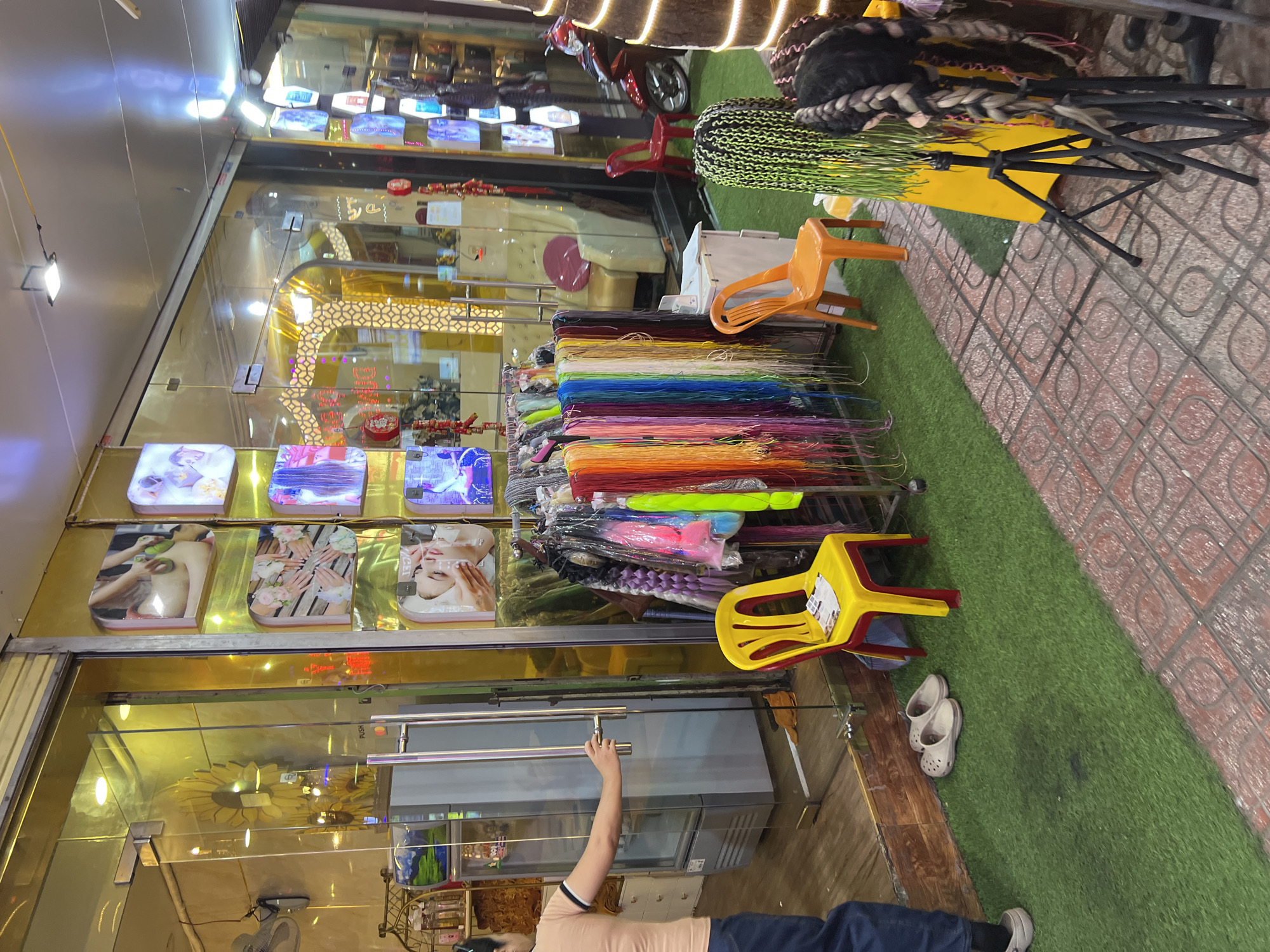 The image depicts the storefront of a hair salon located in the city of Nha Trang, Vietnam, with a focus on hair extension products. The storefront features a large glass door with a metallic handle, and adjacent to it, several illuminated display panels showcase images related to beauty treatments, including massages and manicures. The interior of the salon appears brightly lit with a golden hue, and visible within are salon chairs and a refrigerator.
Outside the salon, a display of vibrantly colored hair extensions hangs from a rack. In front of the salon, artificial green turf covers the ground, on which are placed two simple yellow chairs and an orange chair. A pair of beige slip-on shoes rests on the turf. Further to the right, on the tiled sidewalk, mannequins with elaborate braided hairstyles are displayed, along with tripods. A red motorcycle is parked just inside the salon entrance. The overall scene suggests a retail environment offering hair styling services and products.