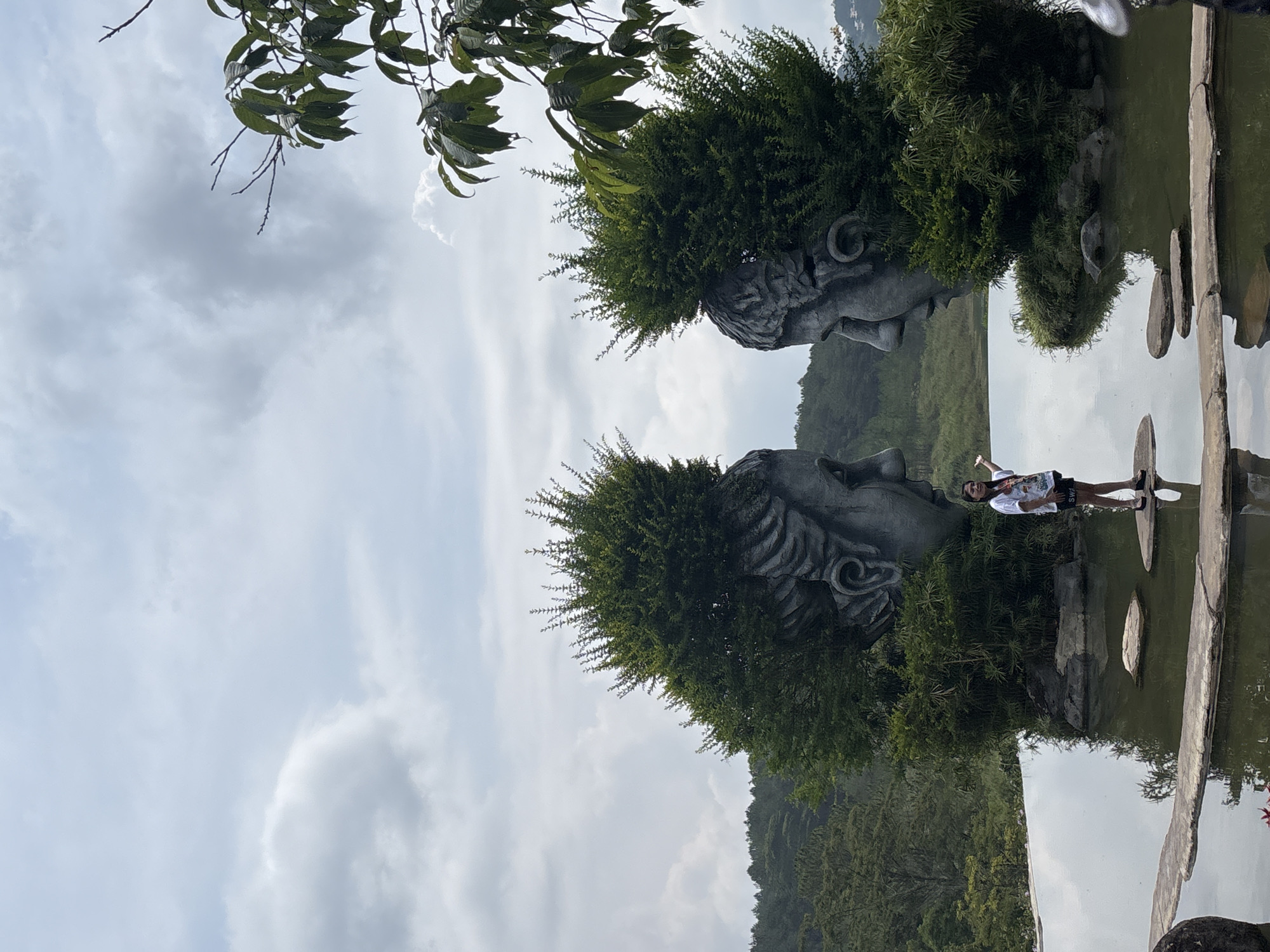 This image captures a scenic view in Da Lat, Vietnam, featuring unique artistic installations and a person posing. The scene is dominated by two large, weathered stone sculptures resembling ancient or mythological heads, positioned on either side of a body of water. Lush green shrubbery forms the "hair" or ornate headdresses for these sculptures, giving them a vibrant and organic appearance.
In the mid-ground, a person, likely a young female, stands on a paved stone pathway or stepping stones that traverse or border the water. She is wearing a white t-shirt with the text "SWA" faintly visible, dark shorts, and sneakers. She is looking towards the camera with a relaxed posture, seemingly posing for a photograph. The water reflects the cloudy sky above and portions of the surrounding landscape and sculptures.
The background reveals rolling green hills or mountains, indicative of a natural, elevated landscape. Overhead, dark green leaves and branches from an unseen tree hang into the frame, partially obscuring the cloudy sky. The sky itself is overcast with diffused light, suggesting daytime with bright, but not direct, sunlight. The overall impression is one of a tranquil and picturesque park or garden setting.