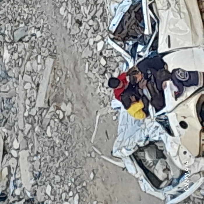 The image depicts a scene in Khan Yunis, Palestinian Territory, characterized by extensive structural damage. The upper two-thirds of the frame are dominated by a vast field of light-colored rubble, consisting of fragmented concrete, stones, and pulverized debris, indicating a severely damaged or destroyed urban environment. The ground beneath this debris appears to be a mix of dirt and smaller fragments.
In the lower third of the image, a heavily damaged white vehicle, consistent with a car or a small utility vehicle, is situated amidst the debris. The vehicle's roof is significantly crushed and distorted, with its front end partially obscured or collapsed. The side panels and doors show visible damage, including large indentations and possible missing components.
Four individuals, appearing to be children, are present around and on the damaged vehicle. One child, wearing a red long-sleeved top and a dark cap, is positioned on the highest part of the crushed roof, facing downwards. To their left, another child in a yellow top is also seated on the roof structure, partially obscured. A third child, dressed in a dark blue or black jacket, is seated lower down within the vehicle's interior or on its chassis, seemingly facing towards the other two children. A fourth child, wearing a dark blue jacket with a white circular emblem on the back, is standing near the driver's side door of the vehicle, with their left hand on the door panel, appearing to interact with or climb onto the vehicle. The children appear to be engaged in exploration or play on and around the wrecked car.