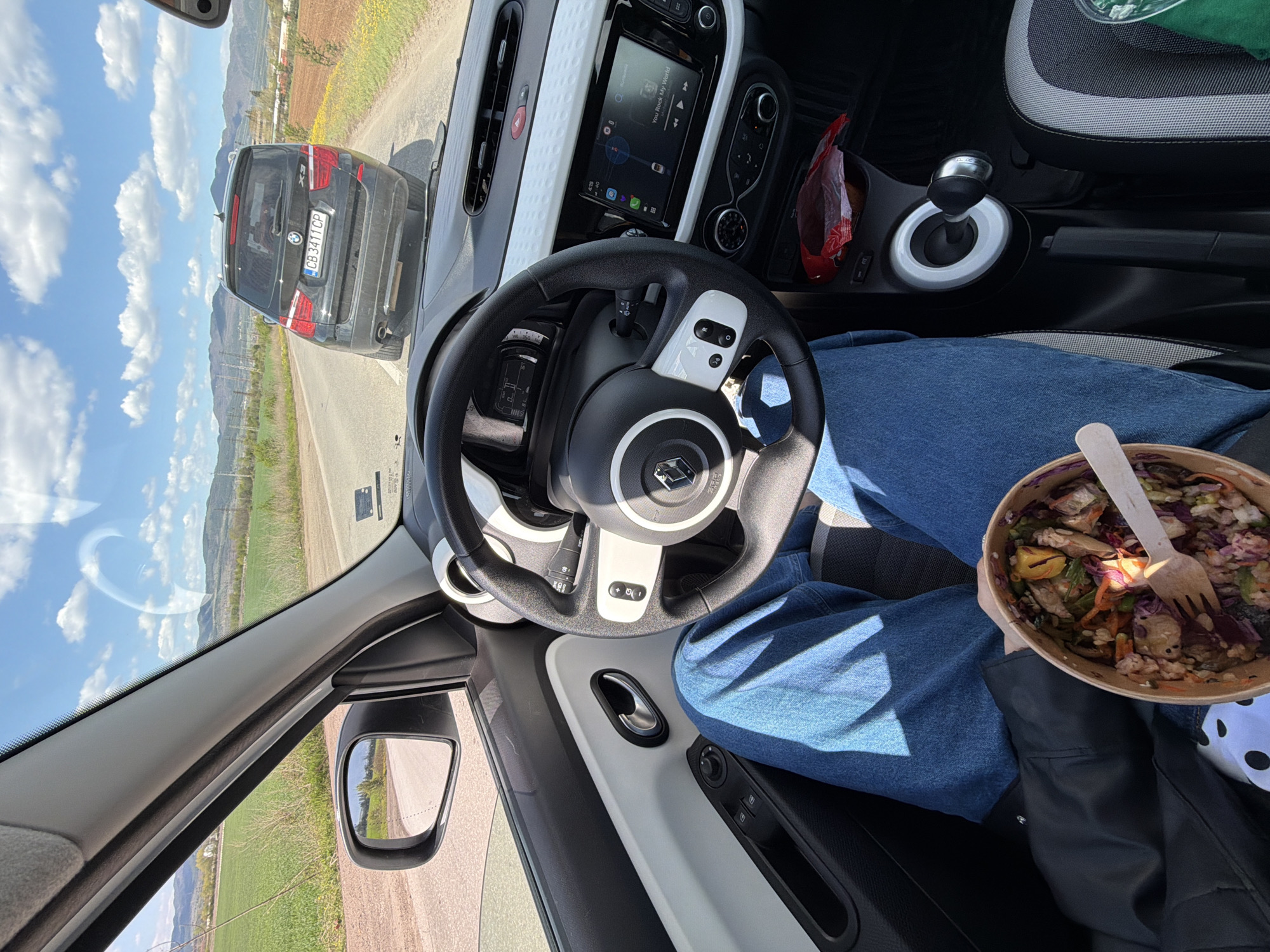 The image captures a first-person perspective from inside a car, looking out through the windshield and into the car's interior. A person's legs, wearing blue jeans, are visible on the right, along with their hand holding a paper container filled with a meal, possibly a salad or stir-fry, with a wooden fork. A black jacket is draped nearby.
Inside the vehicle, a black steering wheel with a prominent Renault logo is central to the foreground. The dashboard behind it features an infotainment screen displaying "You Rock My World," and various controls and air vents are visible. To the right of the person's legs, a gear shift and a red bag are in the console area. The side mirror on the left reflects a glimpse of the road and surrounding landscape.
Outside, the weather is bright and sunny, with a clear blue sky dotted with scattered white clouds. A black BMW X3 SUV is directly in front on a paved road, which has a dirt shoulder. The BMW's license plate, "CB 3411 CP," indicates it is registered in Sofia, Bulgaria. Beyond the road, green fields extend towards distant mountains, suggesting a scenic rural or suburban setting. The scene portrays a person taking a meal break while in a car, likely stopped or moving slowly, in the vicinity of Sofia, Bulgaria.