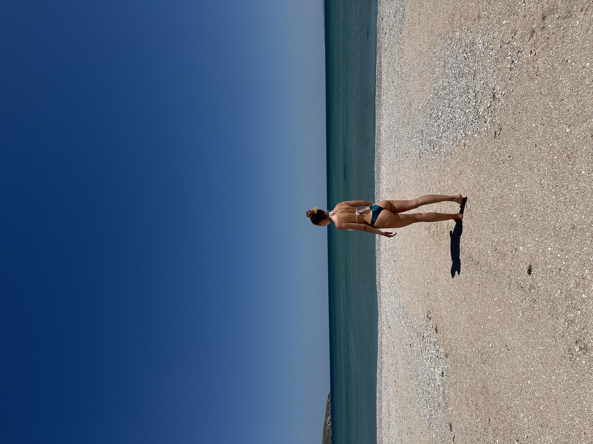 The image captures a person standing on a beach in the city of Unknown, Unknown. The individual, seen from the back, is wearing a two-piece swimsuit. The beach is composed of sand and shells. The ocean meets the beach in a straight line. A shadow of the person extends onto the sand. The sky occupies the left portion of the image and is a clear, unclouded blue.