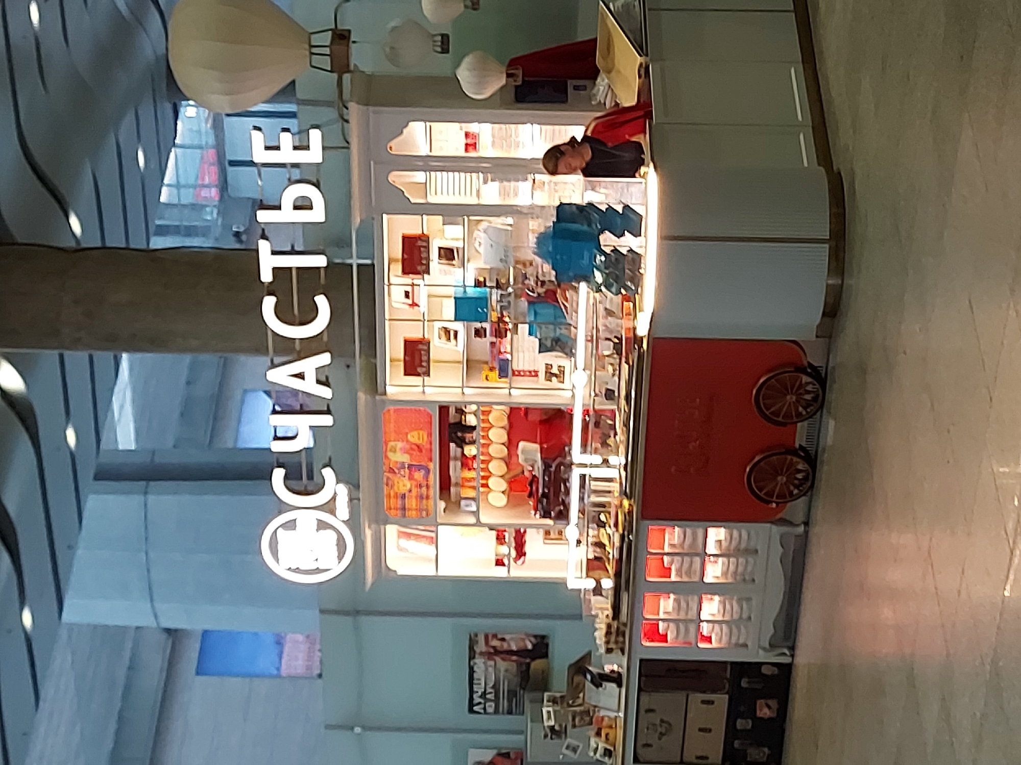 The image shows a kiosk selling confectionery or baked goods, displaying the word "СЧАСТЬЕ" (Happiness) vertically in white letters, an identifying mark of the СЧАСТЬЕ brand, a popular bakery chain located in Saint Petersburg, Russia. The kiosk is predominantly white with red accents and appears to be indoors, possibly within a shopping mall or airport, judging by the architecture visible in the background. A staff member wearing dark clothing is seen behind the counter, likely serving customers. The kiosk displays various items and decorative elements like hot air balloons, adding to its unique visual appeal.