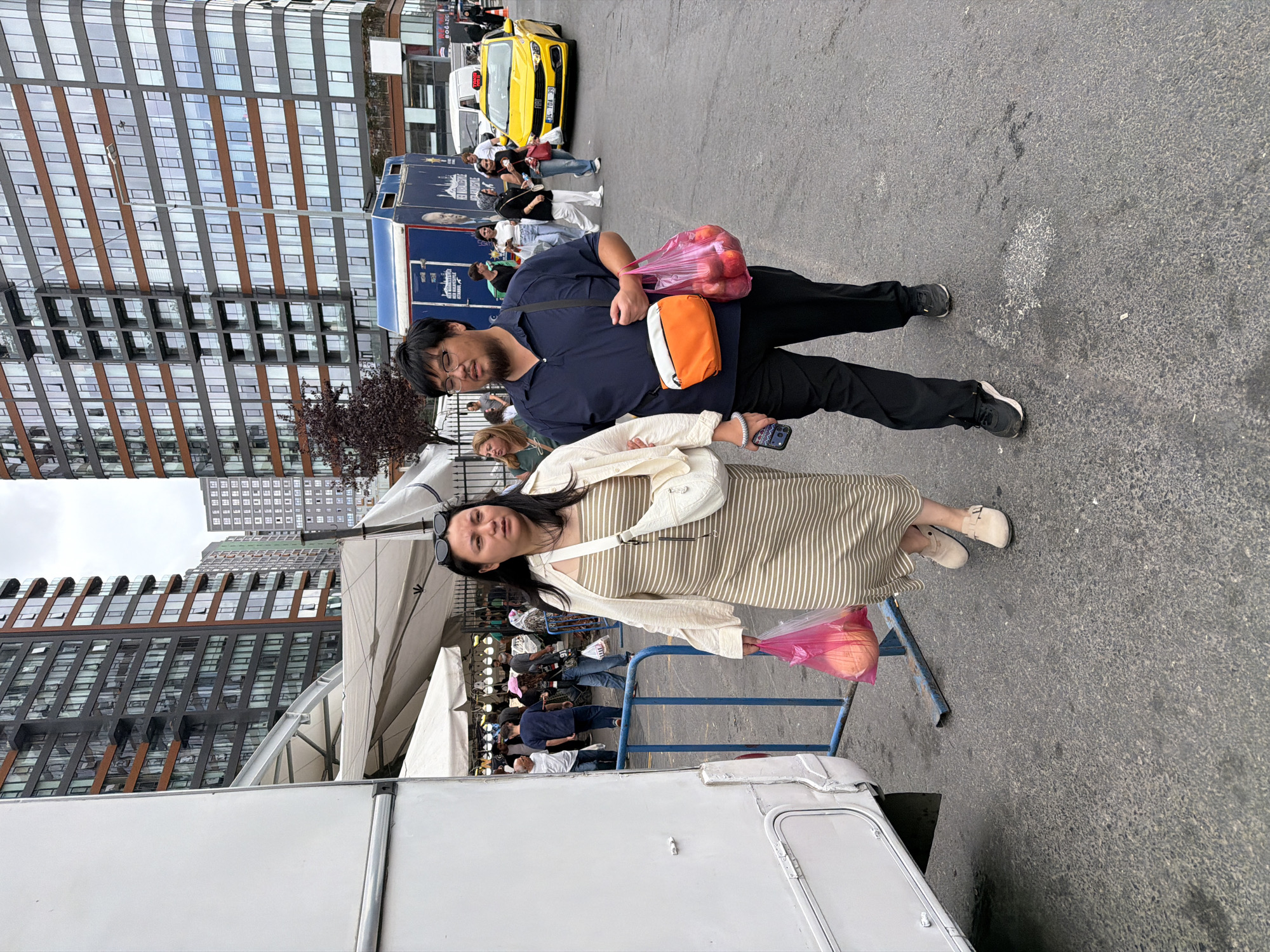 In Kadıköy, Turkey, a man and woman walk together on a street. The woman, wearing a striped dress and an off-white cardigan, carries a pink plastic bag with an orange object inside. The man, in a dark blue polo shirt and black pants, carries a pink plastic bag filled with red fruits, and an orange and white fanny pack. They are in front of a white RV, and in the background, a yellow taxi is visible, along with tall residential buildings and market tents.