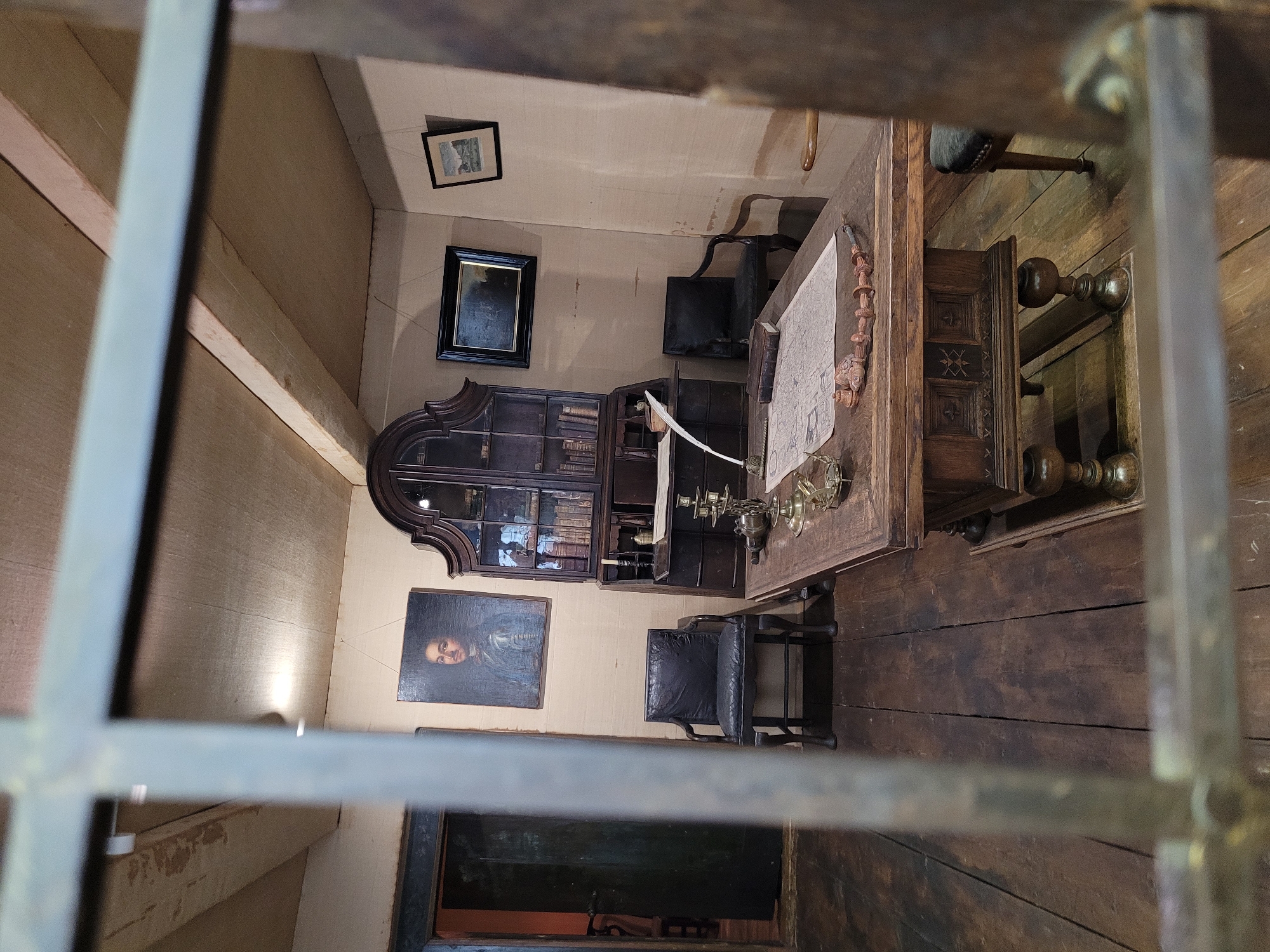 This is an interior shot of a room, likely a study or office, with a wooden floor and ceiling with exposed beams. The room is furnished with antique furniture and objects, suggesting a historical setting. A large wooden desk with intricate carvings is in the foreground, covered with a map, a quill pen, a book, and brass instruments. Two dark leather chairs are placed near the desk and a bookshelf. A portrait of a man in armor hangs on the wall, alongside two framed pictures. A dark wooden cabinet with glass doors, filled with books, stands against the wall. The scene is viewed through what appears to be bars or a fence, indicating the perspective is from outside the room. The overall atmosphere is one of a preserved historical space, possibly a museum exhibit or a recreated historical dwelling. There is no indication that this scene is located in Saint Petersburg, Russia.