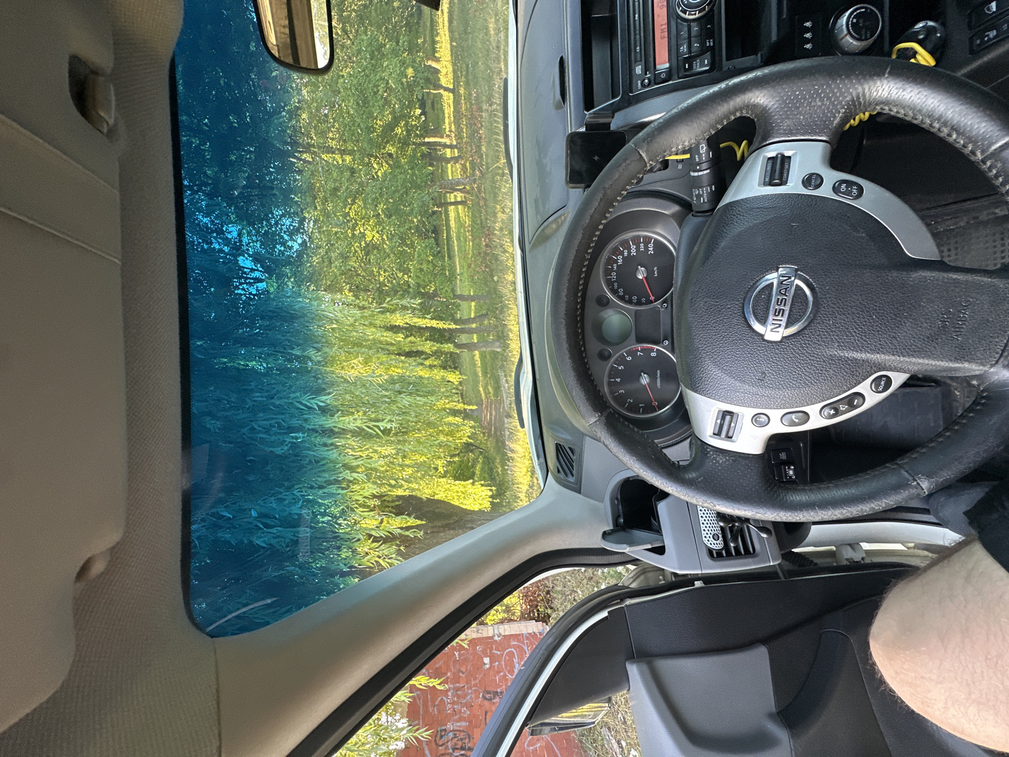 The image shows the interior of a Nissan vehicle, viewed from the driver's seat. The steering wheel, dashboard, and parts of the car's interior are visible. Through the windshield, a park-like setting with trees and sunlight is seen. The blue tint on the windshield suggests it is either a tinted window or has a filter applied. There is no indication of the location being the city of Makiv, Ukraine.