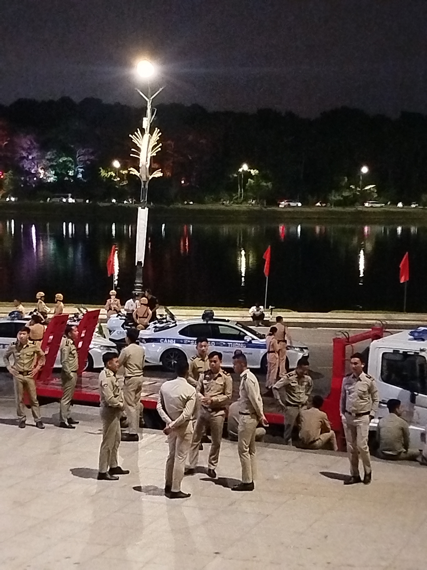 The image depicts a nighttime scene in Da Lat, Vietnam, likely near a body of water. Several individuals in uniform, appearing to be police officers or security personnel, are gathered around marked police vehicles. The officers are mostly wearing light-colored uniforms with dark belts and shoes. Some are standing, while others are seated or leaning against the vehicles.
The environment suggests an outdoor setting at night. A prominent lamppost with decorative elements illuminates the scene, casting light on the pavement and the surrounding area. In the background, a body of water reflects the lights from the city or nearby attractions, creating a shimmering effect. Trees and foliage are visible beyond the water, forming a dark silhouette against the night sky.
The atmosphere appears calm, with the officers seemingly on duty or attending to a situation. There are no obvious signs of distress or urgency. The lighting indicates it is after dark.
Visible text on the police car reads "CSGT" which is an abbreviation for Traffic Police in Vietnam. Another visible portion of text on the police car appears to be in Vietnamese. Two red flags are planted on poles near the police vehicles.
