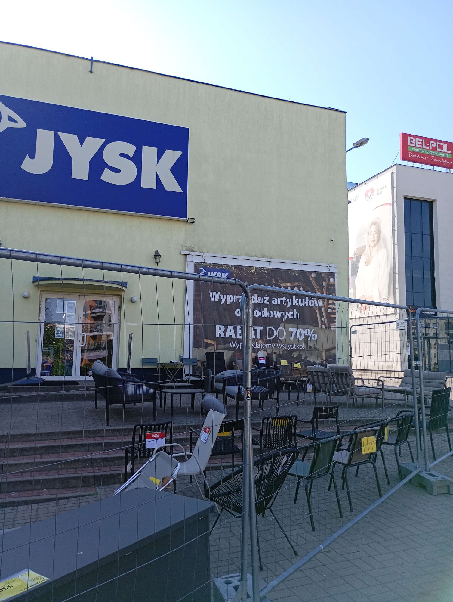 The image shows the exterior of a Jysk store in Białystok, Poland, with outdoor furniture displayed behind a temporary fence. The Jysk logo is prominently displayed in blue on the building's facade. A large banner advertises a sale with "RABAT DO 70%" (up to 70% discount). Various chairs and tables, some with cushions, are arranged on steps and paved areas, partially obscured by the fencing. The scene appears to be on a sunny day. A taller building with a BEL-POL sign is visible in the background.