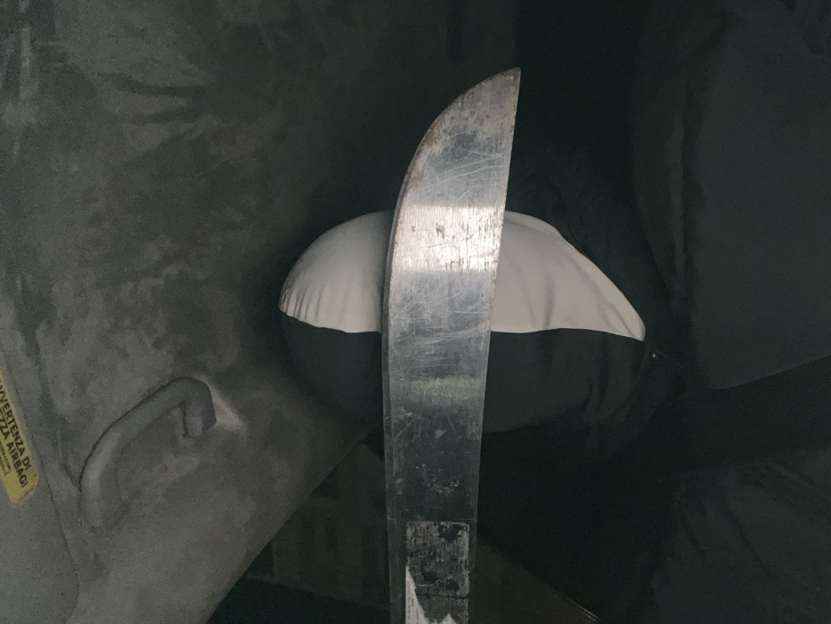 The image displays the interior of a vehicle, specifically depicting a portion of the passenger compartment. A person is visible, primarily showing the upper body and head, obscured by a hood or balaclava featuring a distinct split design of black and white fabric. In the immediate foreground, a large, metallic blade, consistent with a machete or similar long knife, is positioned horizontally across the frame. The blade exhibits signs of wear, including scratches and abrasions, and has a visibly curved tip. Above the individual, the vehicle's grey headliner is visible, along with a grey grab handle and a partial yellow warning sticker, which includes the word "AIRBAG." The surrounding environment is dark. The image itself provides no discernible visual cues or landmarks to confirm the specific city as Plovdiv, Bulgaria.