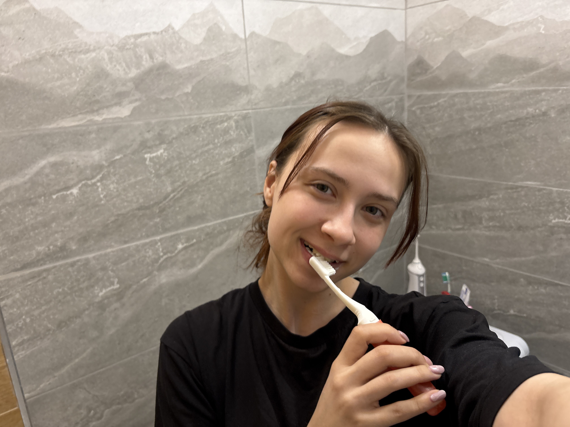 A young woman is taking a selfie while brushing her teeth in a bathroom. The bathroom has light grey tile walls. Toothbrushes and a water flosser are visible on the counter behind her. She is wearing a black t-shirt. It is not possible to confirm the location in the city of Perm, Russia from the visual information in the image.