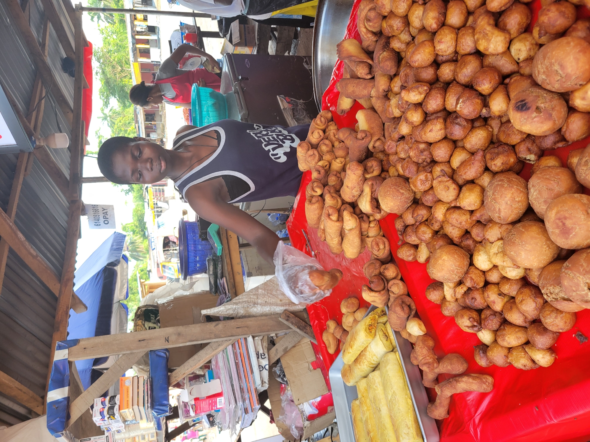A young Black woman, likely in her late teens or early twenties, stands behind a stall laden with fried pastries. She is wearing a dark tank top with a white trim and a graphic print. Her left hand is holding a plastic bag containing one of the pastries. In the background, another woman wearing a red apron is visible.
The stall itself is covered with a red cloth, on which a variety of fried food items are displayed. To the left, a rectangular metal tray holds several long, cylindrical pastries. To the right, a large quantity of smaller, irregularly shaped, golden-brown fried pastries are piled high. Some pastries are coiled or twisted in shape.
The stall appears to be outdoors, with a corrugated metal roof supported by wooden beams overhead. Behind the stall, a glimpse of trees and buildings suggests an urban or semi-urban environment. A blue umbrella is partially visible in the background. Various items are stacked on shelves and on the ground behind the stall, including boxes, books, and plastic bags. The scene is brightly lit, suggesting daytime. The presence of a mobile payment number visible on a sign in the background indicates that transactions are likely being conducted at this market stall. This market stall is located in Egbagu, Nigeria.