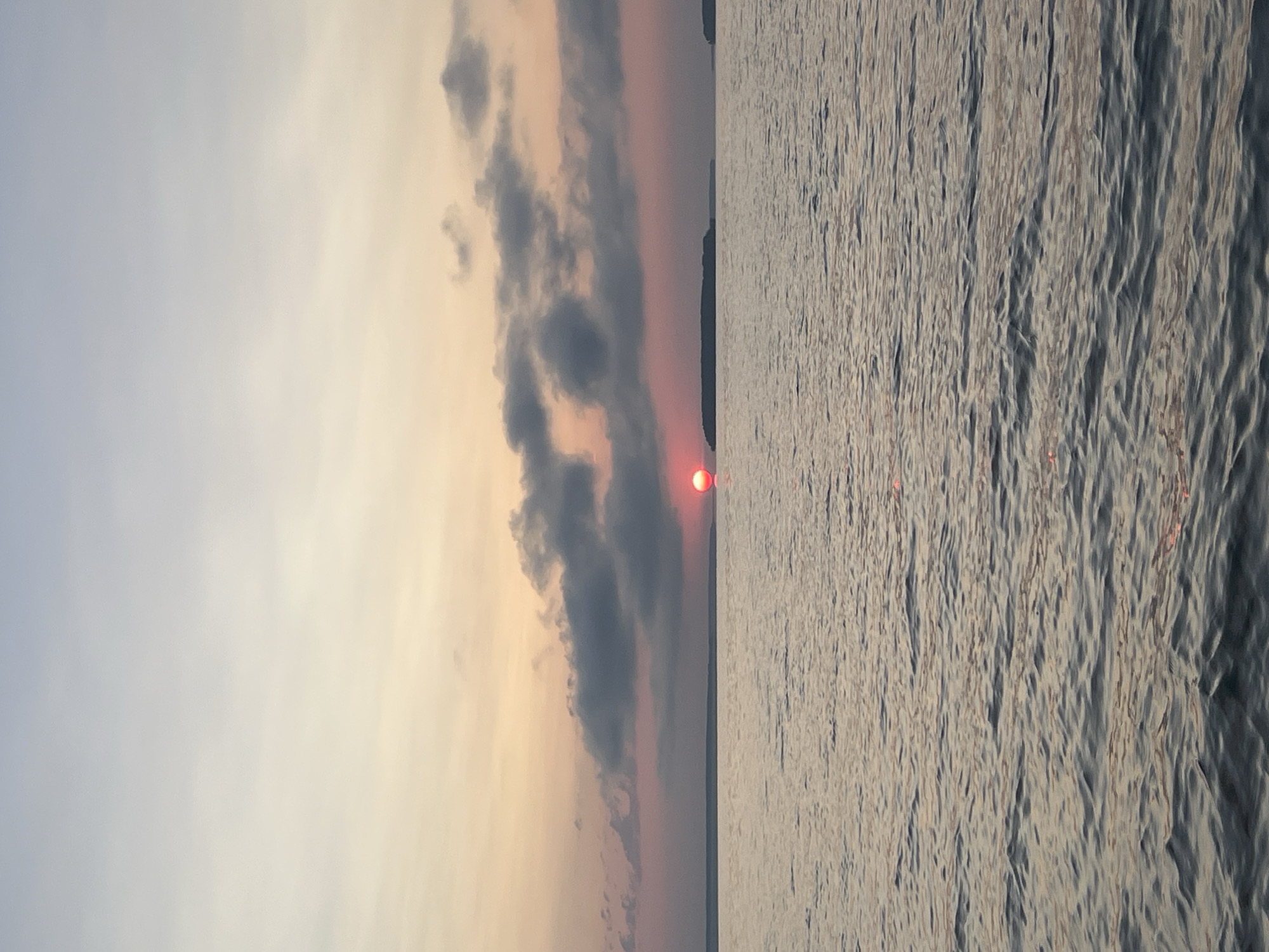 The image depicts a serene sunset over a body of water, possibly a lake or sea, with a distinct red sun partially obscured by dark clouds. The sky transitions from a pale blue at the top to hues of pink and orange near the horizon. Several islands or landmasses are visible in the distance. The water is rippled, reflecting the muted colors of the sky. The scene appears to be located in the vicinity of Novopichugovo, Russia, although this information is not visually derivable from the image itself. There are no people or discernible actions taking place in the photograph.