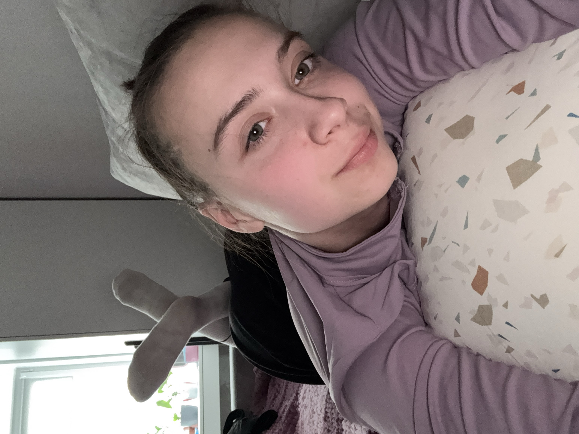 The image is a selfie taken from an overhead perspective, capturing a young woman lying down and looking up at the camera. Due to the image's vertical orientation, her head is positioned towards the left side of the frame. She has fair skin, prominent green-brown eyes, and dark blonde or light brown hair tied back, with a small bun visible. A subtle mole is present above her right eyebrow and another near her nose. Her expression is calm and neutral, featuring a gentle, subtle smile, and her cheeks exhibit a natural flush.
She is dressed in a light purple long-sleeved top, possibly a turtleneck, which appears to be layered over a black garment. Her head rests on a white pillow decorated with a modern, abstract pattern of scattered, irregular shapes in shades of brown, beige, light blue, and grey. Behind her head, a plain light grey surface is visible, while a darker, textured fabric, likely a blanket or throw, is situated below her. To the lower left, a bright window with a white frame indicates it is daytime, providing soft natural light to the indoor setting. A pair of light-colored, plush toy legs are partially visible near the window. The scene portrays a relaxed, private moment within a domestic environment, possibly a bedroom or living space, in Chaikovsky, Russia. There is no visible text.
