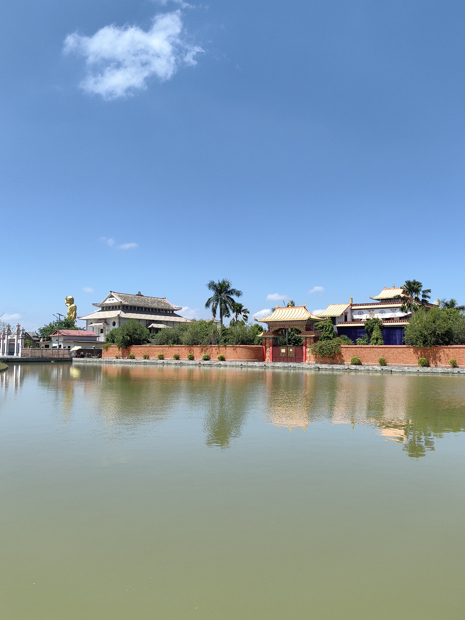 The image shows a tranquil scene with a body of water in the foreground, reflecting the buildings and sky. In the background, there are several structures, seemingly part of a temple or monastery complex, with traditional architecture featuring golden roofs and intricate designs. A large golden statue of a figure is visible to the left, standing tall against the blue sky. The buildings are surrounded by a red brick wall and lush green trees. The sky is clear blue with a few scattered white clouds. The overall impression is one of peace and serenity, typical of a religious or spiritual site. The presence of traditional architecture, the golden statue, and the serene atmosphere suggests a location of cultural and religious significance. Given the context of Lumbini Bazaar, Nepal, this could be a part of the Maya Devi Temple complex or a nearby Buddhist monastery.