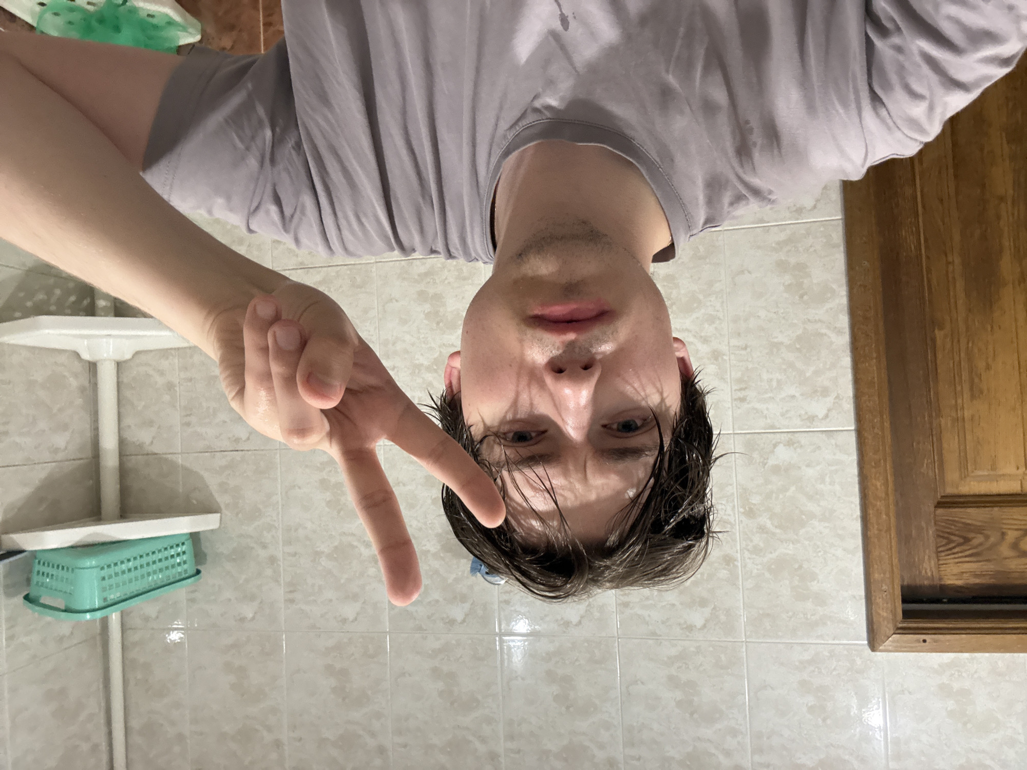 A young Caucasian male with damp, dark hair and a light stubble is taking a selfie in a tiled bathroom. He is wearing a grey t-shirt and is holding up his right hand in a peace sign. Behind him, the wall is covered in light-colored, textured tiles. To his left, a portion of a wooden door is visible. To his right, a white shelf with a teal plastic basket is mounted on a white pole. The overall lighting suggests indoor illumination.