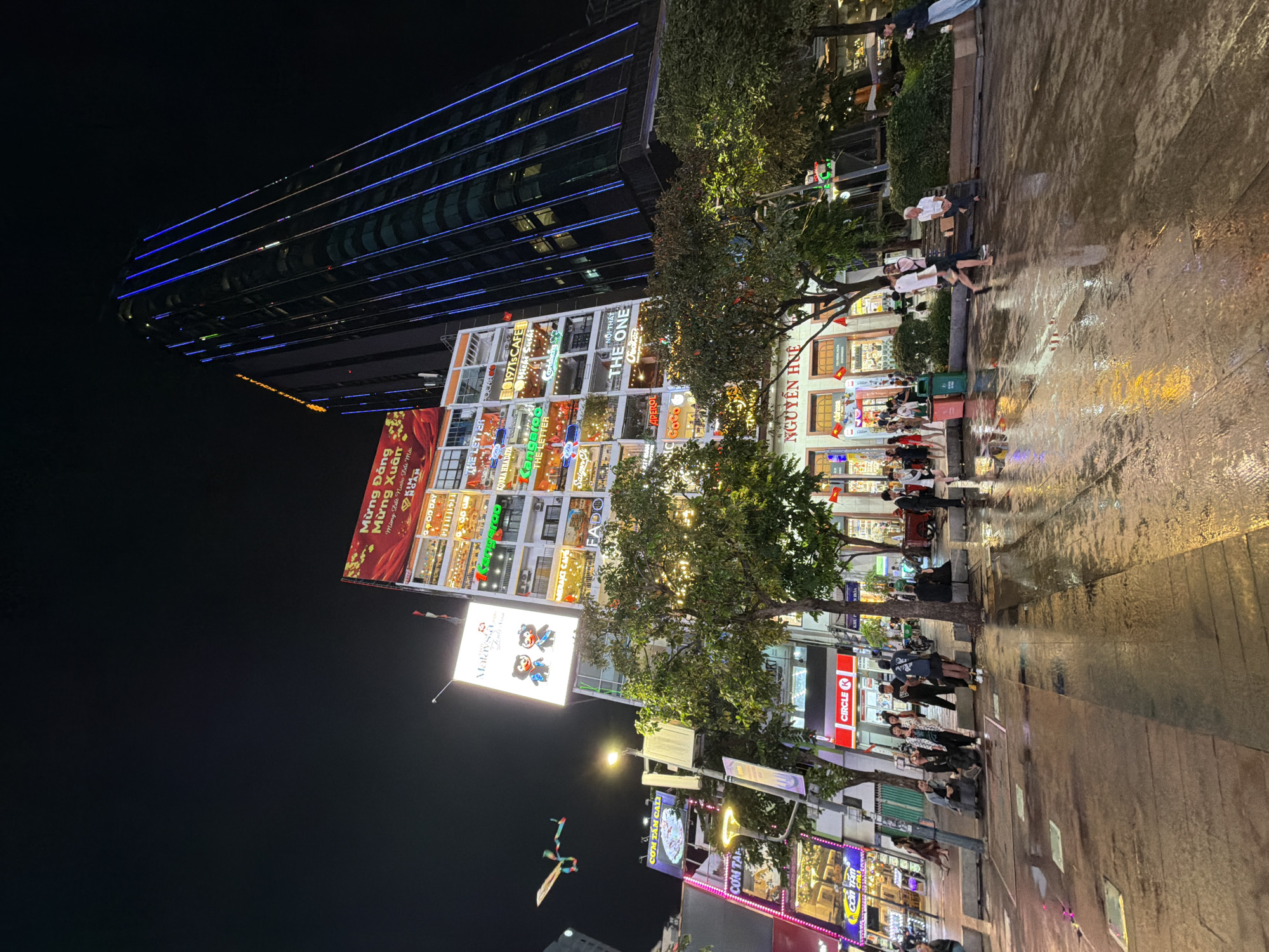 Captured at night in Thủ Đức, Vietnam, the image features a street scene juxtaposed with modern architecture. A tall, dark building with horizontal blue neon lights stands adjacent to a brightly lit building with a large advertising banner. Trees flank the street, and a mix of pedestrians populate the sidewalks. Storefronts with colorful signs and advertisements line the street. The scene is wet, with reflections visible on the ground.
