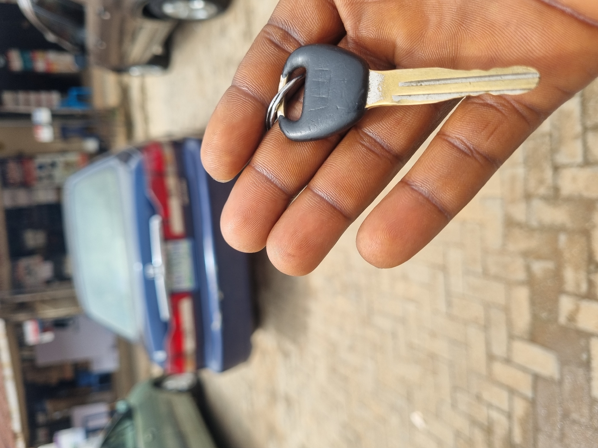 An adult dark-skinned hand is holding a metal car key with a black plastic head and metal ring on the index and middle fingers. A dark blue car is in the background, parked on a beige brick paved surface in an urban setting. The exact location in Jos, Nigeria is not identifiable from the image. Other blurred cars and buildings are also in the background.