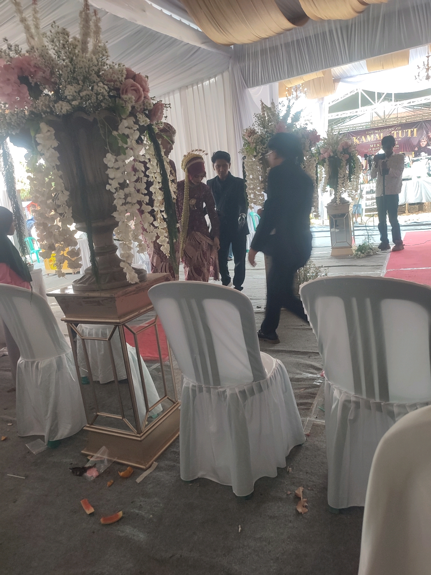 The image depicts an outdoor wedding reception venue, likely in Kecamatan Haurgeulis, Indonesia, judging by the presence of a banner with text in Indonesian. The setting appears to be under a large tent with white fabric draping, and a golden-colored canopy. In the foreground, several white-covered chairs are arranged, some of which are partially obscured by the angle of the camera. The ground is a concrete or asphalt surface, with some scattered debris, including pieces of fruit and plastic wrappers.
In the mid-ground, a large floral arrangement in a dark urn is prominently displayed on a golden stand. The flowers are a mix of white, pink, and green. To the right of the floral arrangement, a man in a white long-sleeved shirt and dark pants is standing and holding a microphone, suggesting he might be an MC or performer. Behind him, there is a stage or raised platform with more floral decorations and possibly other equipment.
Further to the left, a bride and groom are visible. The bride is wearing a traditional maroon outfit and a headdress. The groom is dressed in a dark suit. They appear to be the focal point of the event. Several other individuals are present in the background, including a person in a dark suit standing to the left of the bride and groom, and another person in a dark jacket walking away from the camera. The overall atmosphere suggests a formal celebratory event. The lighting appears to be natural and possibly supplemented by artificial lighting, though none are directly visible.