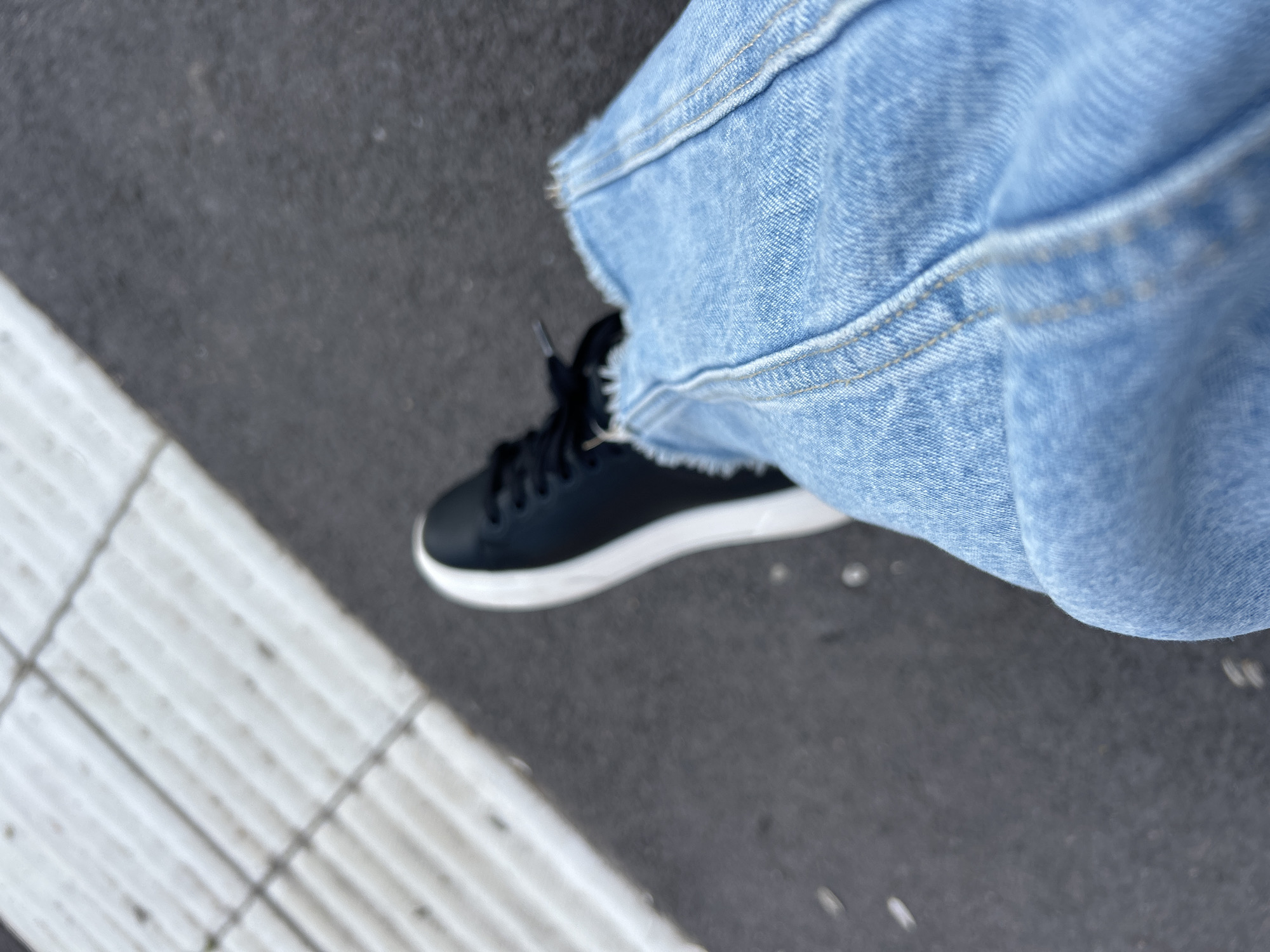 An outdoor, low-angle, eye-level shot shows a person's legs and feet. The left foot is wearing a black low-top sneaker with a white sole. The right leg is clad in light-wash denim jeans with a raw, frayed hem. The ground beneath is asphalt, with a textured white surface in the upper left quadrant, possibly tactile paving in Helsingborg, Sweden, indicating a pedestrian area or transit stop. The lighting suggests it is daytime.