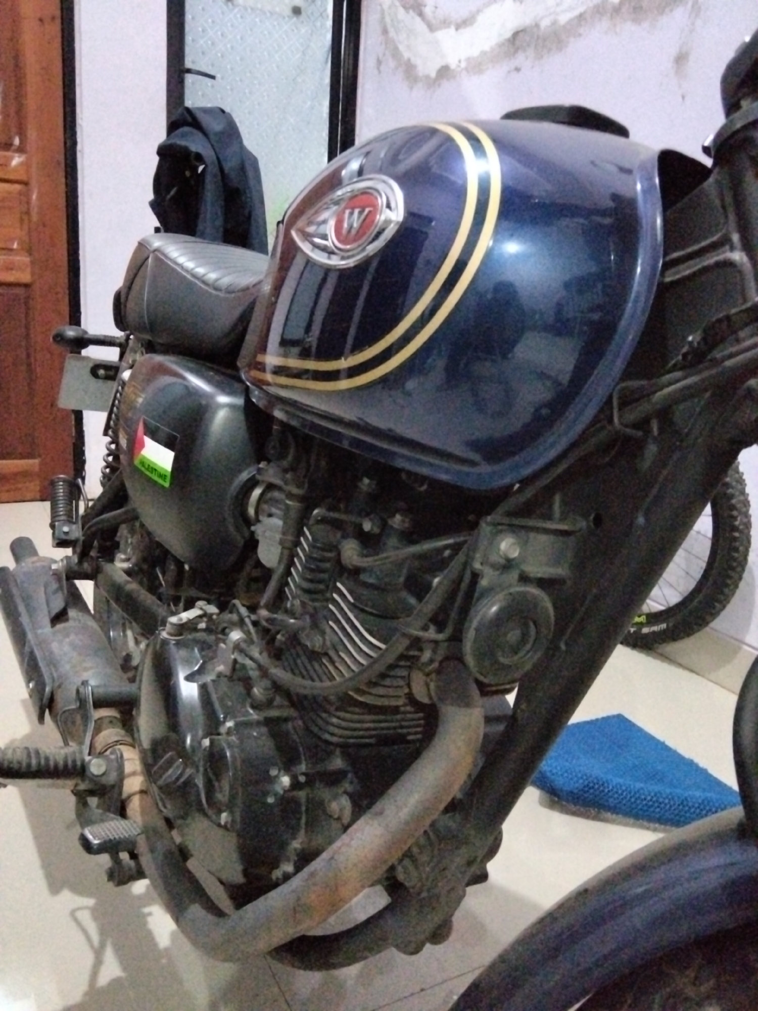 The image shows a dark blue motorcycle parked indoors, possibly in a garage or a residential area. The motorcycle has a distinct emblem on its fuel tank, featuring a "W" inside a circular design. A sticker of the Palestinian flag with the word "PALESTINE" is visible on the side of the motorcycle. A black jacket is draped over the seat. The background shows a tiled wall and wooden elements, suggesting an indoor setting. The flooring appears to be tiled. There is a blue object, possibly a cloth or mat, on the floor near the motorcycle. The overall lighting is dim, with some light sources illuminating the scene. No people are visible in the image. The location is identified as Rawa Atug, Indonesia, though this information is not visually ascertainable from the image itself.