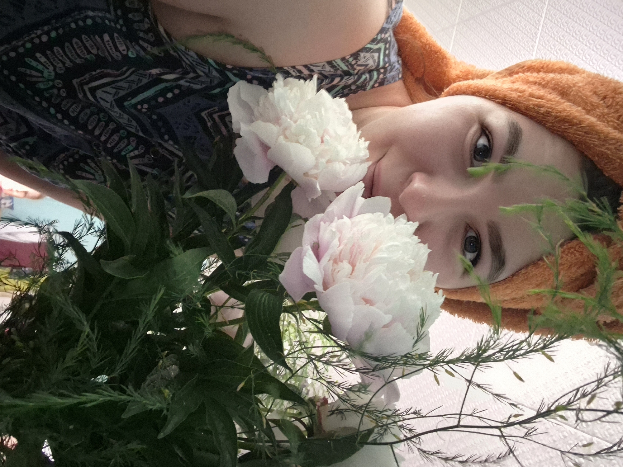 A close-up, medium shot captures a young woman with a bright orange towel wrapped around her head, obscuring her hair. She is looking into the camera, with delicate white peonies in the foreground, partially covering her face. Her left eye is clearly visible, as is her nose and mouth. She wears a dark blue and teal patterned dress with a geometric design. The background consists of a textured wall and some greenery from the flowers. The image is shot indoors, likely in a residential setting, and there is no indication of the specific city of Kharnavy, Russia, in the scene itself.