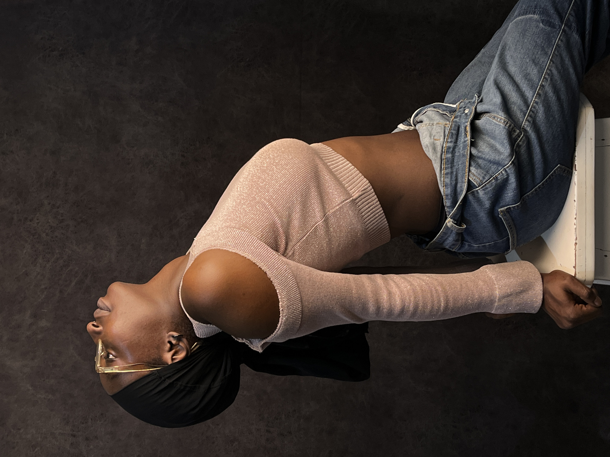A person with brown skin is reclining upside down against a dark background. The person is wearing a light pink long-sleeved top and blue jeans that are unbuttoned. They are wearing gold-rimmed glasses and a black head covering. They are sitting on a white object with their arms outstretched and holding onto the object for support. The location of the image is not known and cannot be determined from the visual data.