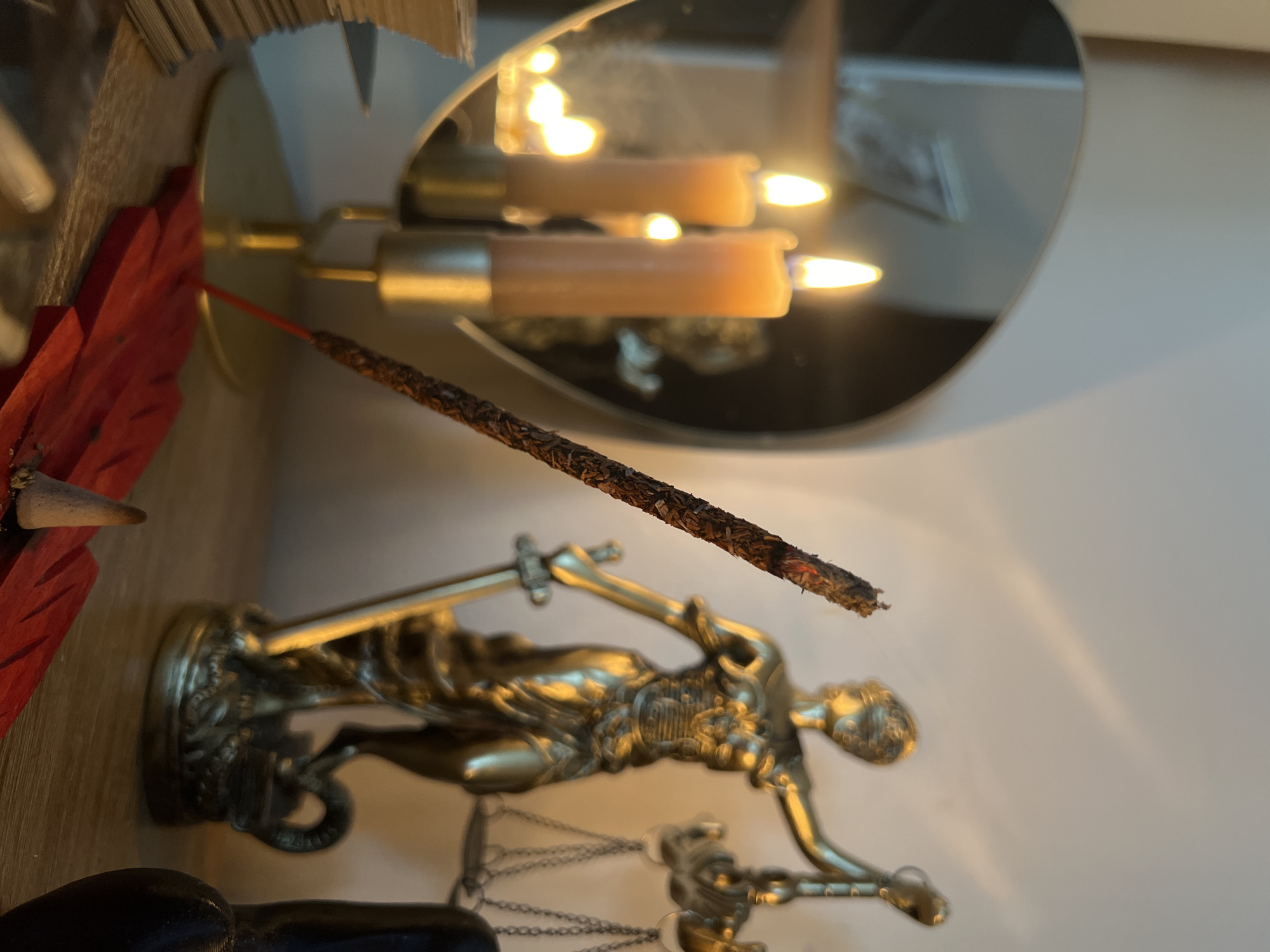 The image depicts a dimly lit, indoor scene. In the foreground, a burning incense stick rests on a red, leaf-shaped holder. To the right, a golden statue of Lady Justice, holding a sword and scales, stands on a wooden surface. In the background, two lit candles in brass holders are reflected in a heart-shaped mirror. The overall ambiance suggests a ritualistic or spiritual setting. There is no visible indication of the location being in Tychy, Poland.