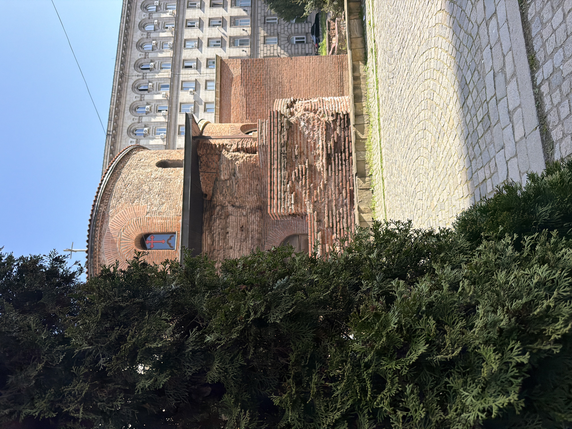 An outdoor, eye-level photograph captures a scene in Sofia, Bulgaria, featuring ancient architecture juxtaposed with a more modern building, partially framed by dense greenery.
In the foreground, a vibrant mass of dark green evergreen bushes fills the lower part of the image, slightly obscuring the structures behind them.
In the middle ground, two distinct historical brick structures are visible. On the left, a rounded, ancient building, likely a church or rotunda, constructed from varied reddish-brown bricks. Its curved wall features a small rectangular window with a distinctive red cross on a blue background. Adjacent to this, to the right, are weathered brick ruins, showcasing exposed and eroded brickwork, hinting at a long history. To the far right, a light-colored cobblestone path with patches of green moss or grass runs alongside these ruins.
The background is dominated by a tall, multi-story building, likely from the early 20th century or later, constructed from light grey stone or concrete. It features numerous symmetrically placed arched windows, many of which have modern air conditioning units installed beneath them. Architectural details like cornices and decorative elements are visible along its facade. The building rises against a clear, bright blue sky, which spans the upper left portion of the image. A thin, dark cable or wire is faintly visible stretching across the sky.
The scene depicts a sunny day, with no people visible and no specific activity taking place. There is no discernible text in the image.