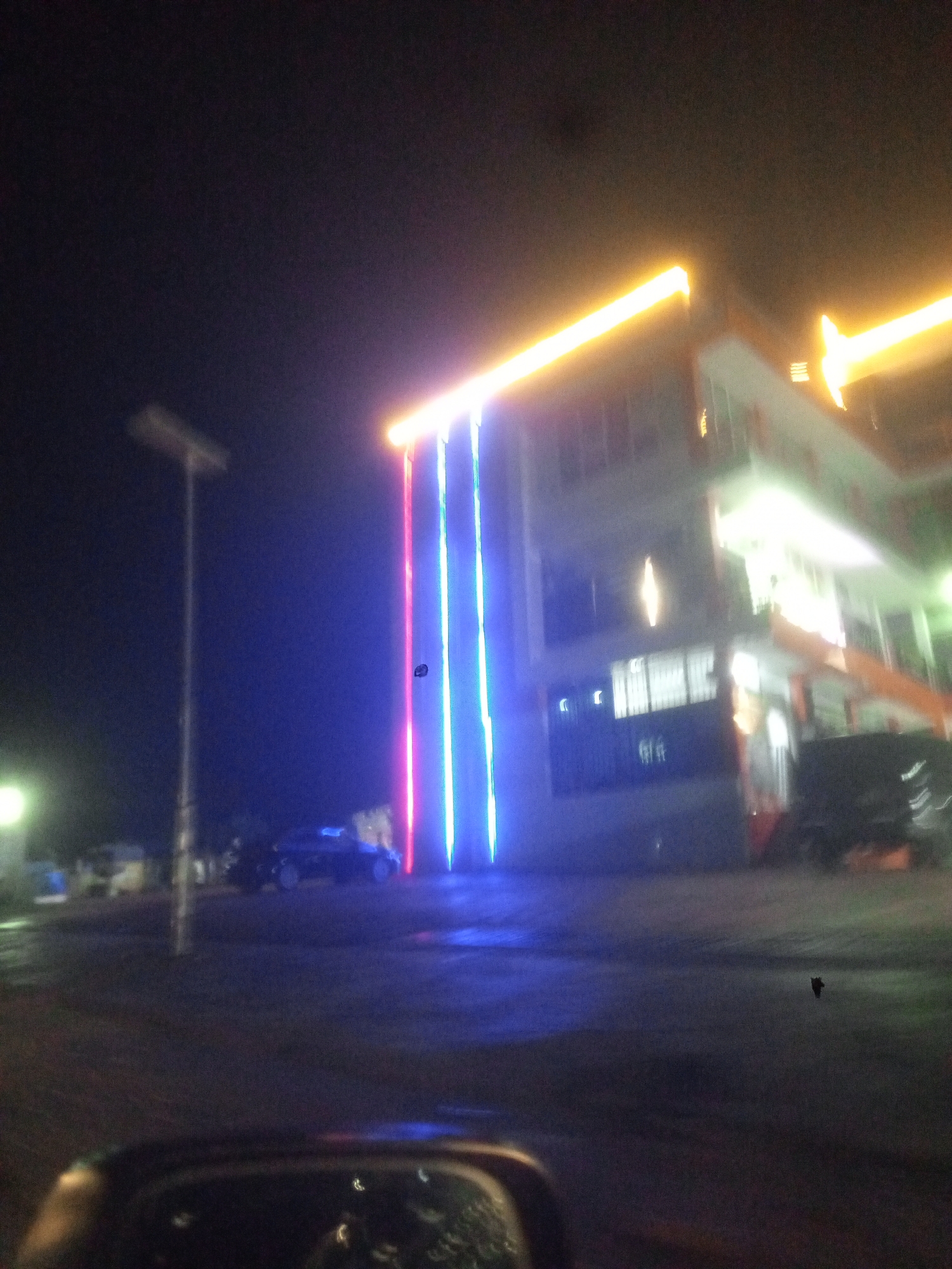 At night in Gwarinpa, Nigeria, a multistory building stands illuminated with yellow lights along its roofline and three vertical stripes of red, white, and blue lights descending from the roof. A tall street lamp stands to the left of the building. A dark car is parked on the wet ground in front of the building. The image is taken from inside a car, with a portion of the car's interior visible at the bottom. The overall scene has a strong blue tint.