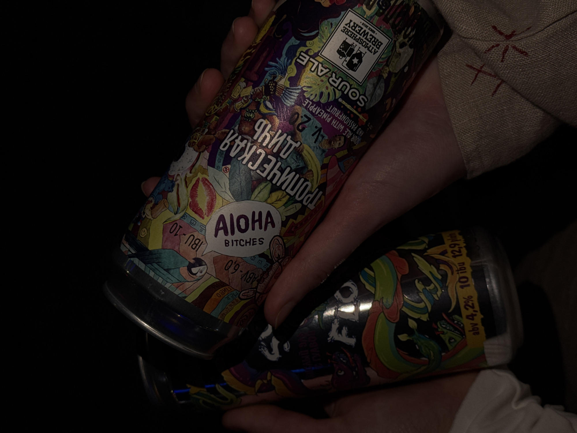 A hand holds two cans of Atmosphere Brewery sour ale. The top can is labeled "Tropical Day V.2.0 Sour Ale with Pineapple and Passionfruit." The labels also show that the brewery was founded in 2016. The ale has an ABV of 4.2% and contains 10 IBU in 12.9 degrees of beer. The other can is labelled "Aloha Bitches", with ABV 6% and 10 IBU. The picture was taken in Perm, Russia.