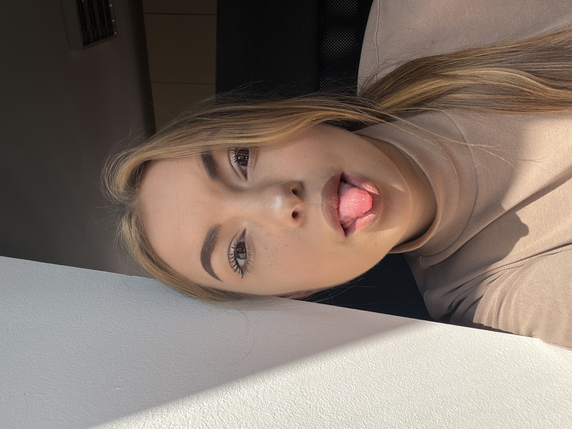 A young woman is depicted in a waist-up indoor portrait, presumed to be located in Sofia, Bulgaria. She has light skin, dark eyebrows, eyeliner, and long, multi-toned blonde and light brown hair pulled back into a ponytail, with some strands loose around her face. She is making a playful expression by sticking out her pink tongue. She wears a light beige long-sleeved turtleneck top. Intense natural light, likely sunlight, originates from the left, casting a sharp diagonal shadow across the textured white wall behind her and illuminating the left side of her face and garment. The right side of her face, shoulder, and the background are in deeper shadow. Visible in the shadowed background are the dark back of a chair, dark wooden cabinetry or shelving, and an overhead air conditioning vent or similar fixture.