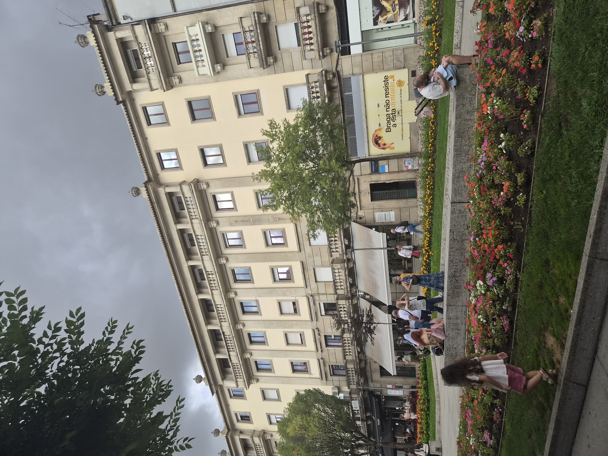 In Braga, Portugal, a man sits on a concrete ledge in front of a flowerbed, holding a baby. A young girl walks in the foreground, facing away from the camera. Behind them, a wide street is lined with shops and a pale yellow building with balconies. Several people are walking along the sidewalk and seated at outdoor cafe tables. The sky is overcast.