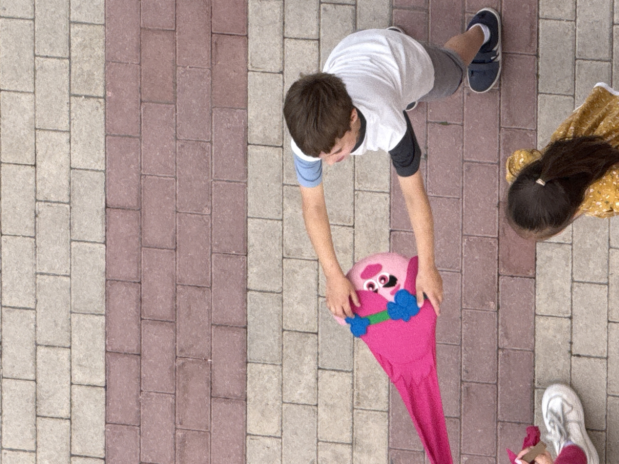 An overhead shot shows two children interacting with a large, pink, stuffed animal that resembles a character from a children's show, possibly a troll. The stuffed animal is elongated and has a cartoonish face with large eyes. It is positioned horizontally in the frame. A boy wearing a white t-shirt with blue and black sleeves and grey shorts is on the right side of the image, holding the stuffed animal with his right hand and touching its head with his left. A young girl wearing a yellow floral dress is on the bottom right of the frame, facing away from the camera. Her dark hair is pulled back into a ponytail. Another child's foot wearing a white sneaker is visible on the bottom left of the frame. They are all standing on a pathway paved with rectangular bricks in alternating colors of tan and reddish-brown. There are no discernible landmarks to indicate a specific location within Moscow, Russia.