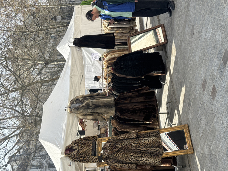 An outdoor market scene is depicted under clear daylight on cobblestone pavement in the city of Unknown, Unknown, consistent with a French urban environment. Multiple white canopy tents are erected, displaying numerous fur coats on metal racks. Coats vary in color and pattern, including leopard print, brown, black, and grey/tan. Two framed mirrors, one gold and one brown, lean against the ground near the coat displays. A partial French sign visible under a tent reads "DESTOCKAGE FOURRURE VINTAGE" (Vintage Fur Clearance). In the foreground right, an adult male, wearing a blue, green, and black jacket over a light blue shirt, dark trousers, and dark shoes, stands facing left, engrossed with a mobile phone held in his hands. In the background, partially visible under a tent, an adult female with dark hair is situated behind a counter, with another individual's head visible further back. Bare-branched deciduous trees and multi-story stone buildings with traditional architecture line the background, accompanied by a traditional lamppost.