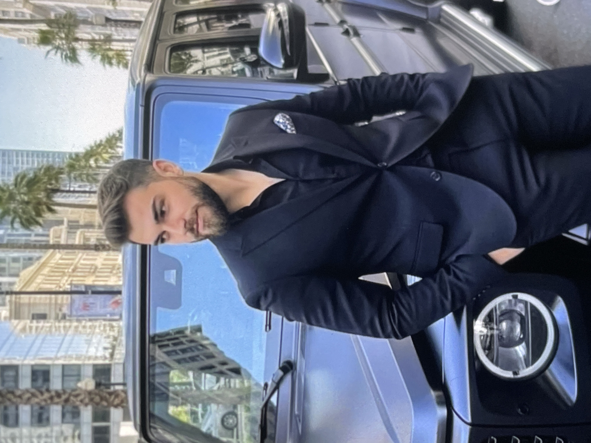 A man is standing next to a black SUV in a city. The man is wearing a black suit and a black shirt. He has short brown hair and a beard. He is looking to his left with his hands in his pockets. The SUV is parked on the street in front of a building. There are palm trees and other buildings in the background. The sky is blue.