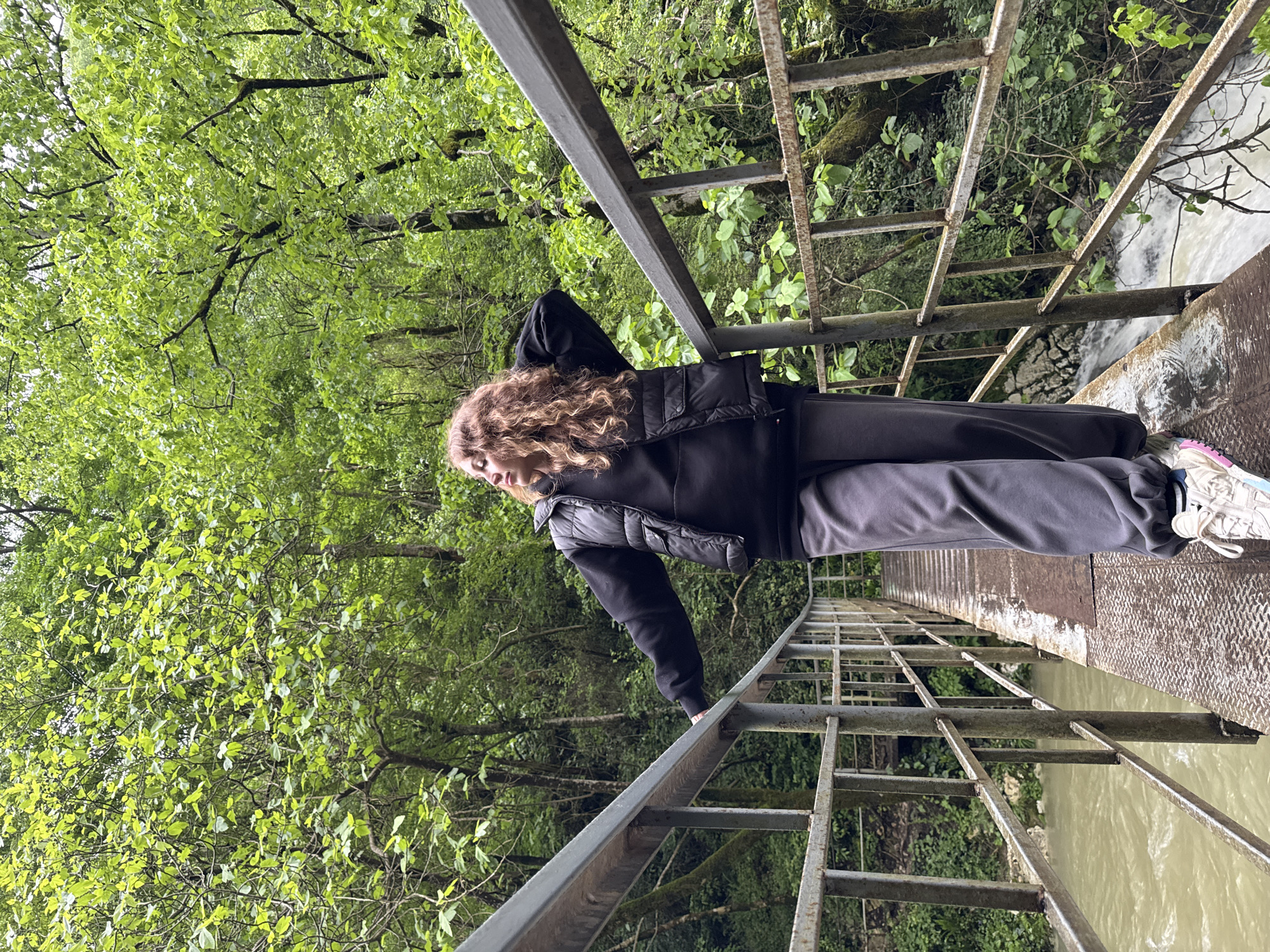A young woman stands on a narrow, metal bridge overlooking a body of water in Tsabal, Abkhazia. She wears a black jacket and gray sweatpants. The bridge is constructed of metal beams and a textured metal walking surface. The surrounding environment consists of lush green trees and foliage. The water is murky and reflects the surrounding greenery. The photograph is taken from a high angle, looking down at the woman and the bridge.