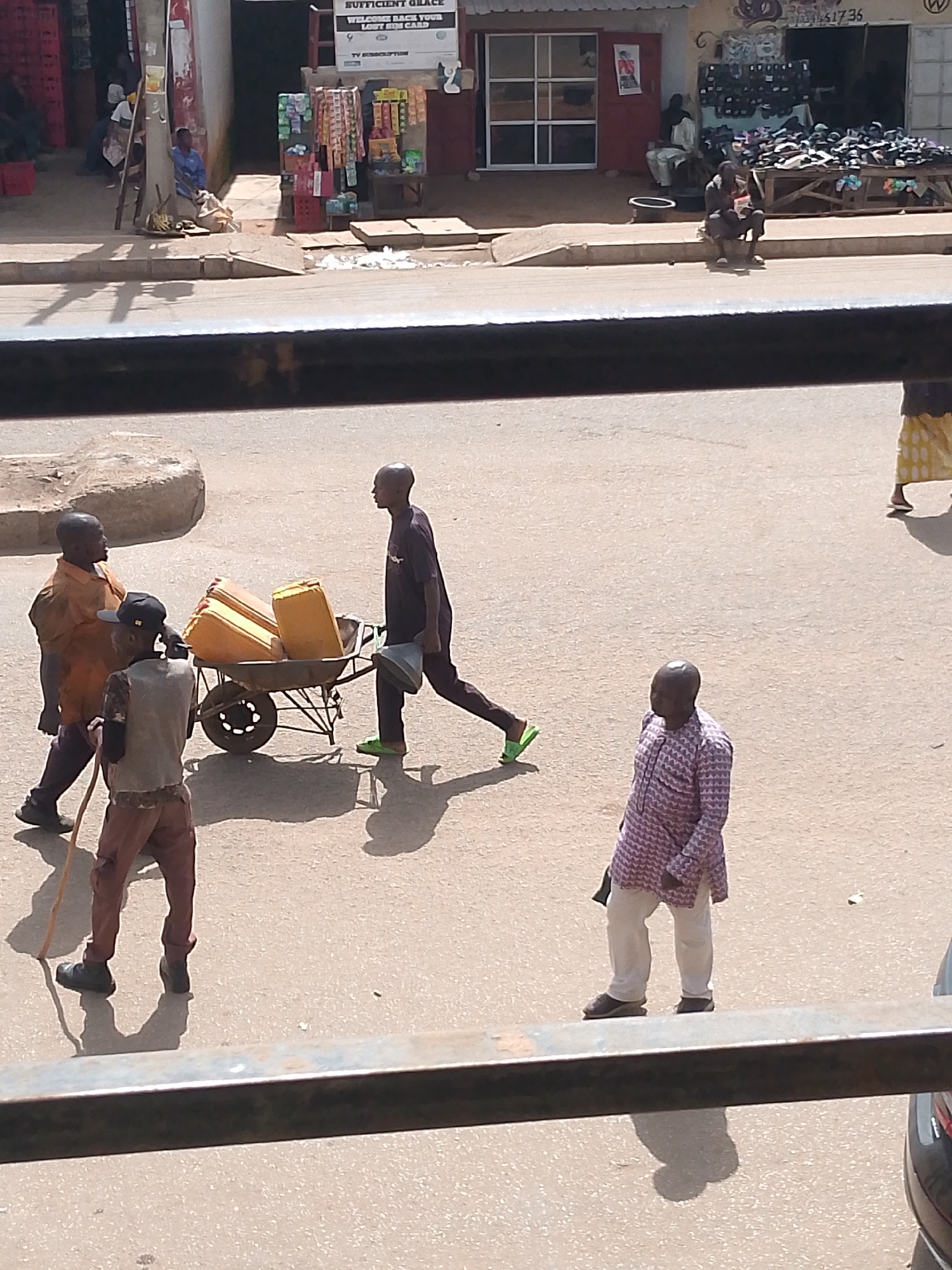 This media file captures a street scene in Bukuru, Nigeria, under bright daylight, suggesting a warm day. In the foreground, a person is walking with a stick. Further back, three men are actively engaged. Two men are near a wheelbarrow filled with several large, yellow jerrycans, likely containing water. One of these men appears to be pushing or assisting with the wheelbarrow, while the other stands beside it, possibly discussing their task. A third man, dressed in a dark t-shirt, walks away from the wheelbarrow carrying a grey bucket and wearing bright green sandals. In the background, a woman in a patterned skirt walks away from the camera. Another man in a patterned tunic and light-colored trousers stands observing the scene. The setting appears to be a paved street in front of shops. One shop has a sign with the text "SUFFICIENT GRACE WELCOME BACK YOUR LOST SIM CARD" and "TV SUBSCRIPTION". The overall impression is of a bustling, everyday scene with people carrying out their daily activities in a commercial area.