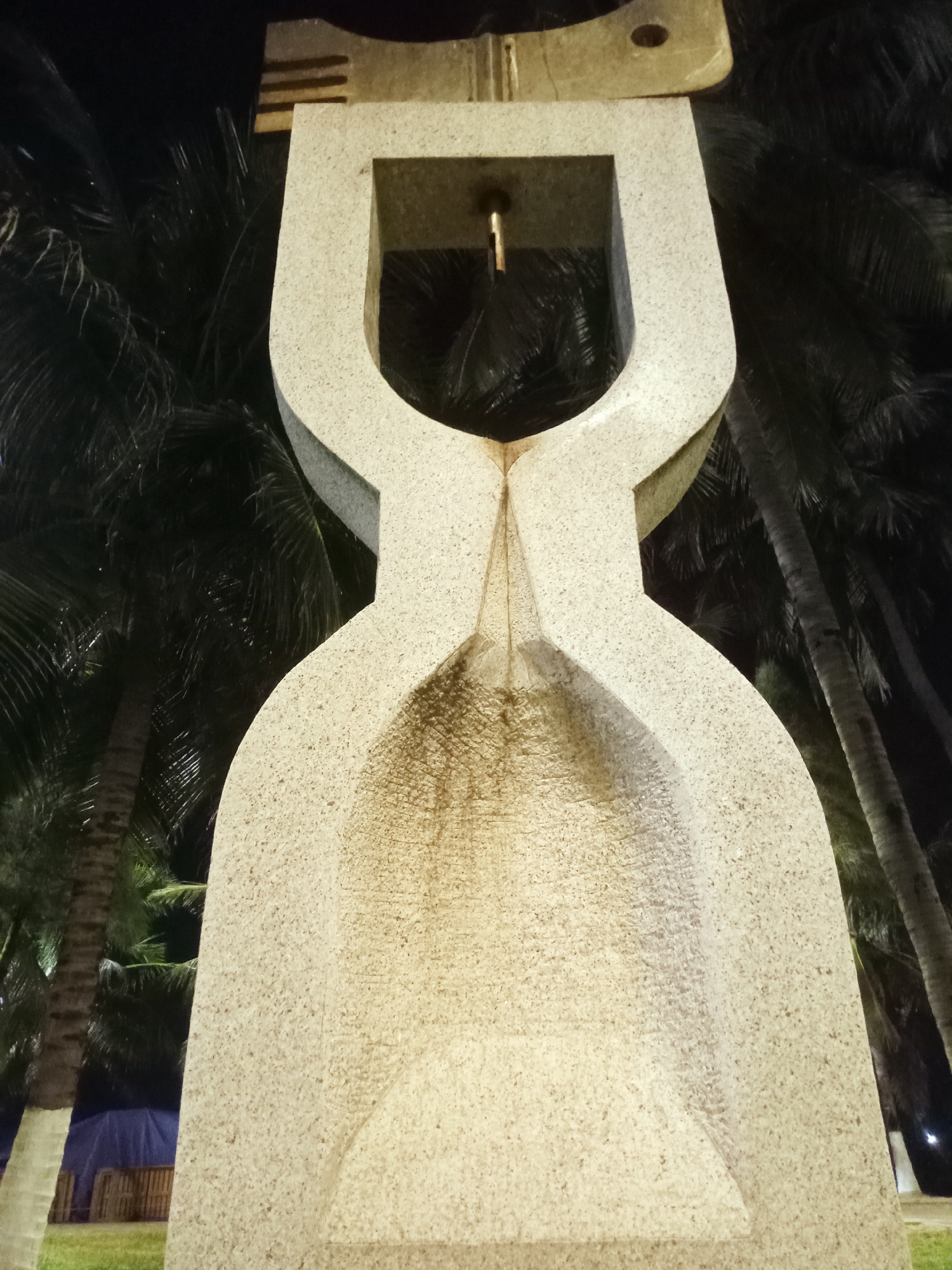 A low-angle shot reveals a stone monument, possibly a water fountain, with a distinctive hourglass shape dominating the frame. The monument is constructed from light-colored, rough-textured stone, displaying visible weathering or staining along its concave surfaces. At the top of the structure is a rectangular element with horizontal striations and a rounded, key-like protrusion, with a small pipe extending down into the upper open area. Dark vegetation, particularly palm trees, is visible in the background, creating a stark contrast against the illuminated stone. The lighting suggests a night scene, with the monument being artificially lit. At the bottom of the image, there is a glimpse of a green grassy area and a blue and white structure or tent. This image was likely taken in Nha Trang Ward, Vietnam.