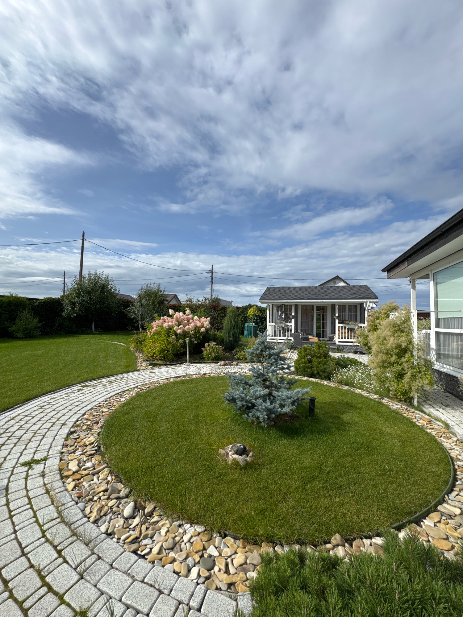 The image depicts a well-maintained garden and yard with a small, white wooden house with a dark roof in the background. A paved pathway, made of light-colored rectangular stones, curves through the green grass. A circular garden bed with a small blue spruce tree in the center is surrounded by river stones. A cluster of pink hydrangeas is visible to the left of the house. The environment suggests a suburban or rural setting. The sky is partly cloudy with white clouds against a blue background. The image is taken from a ground-level perspective, looking slightly upwards. The location is identified as Oliki, Russia.