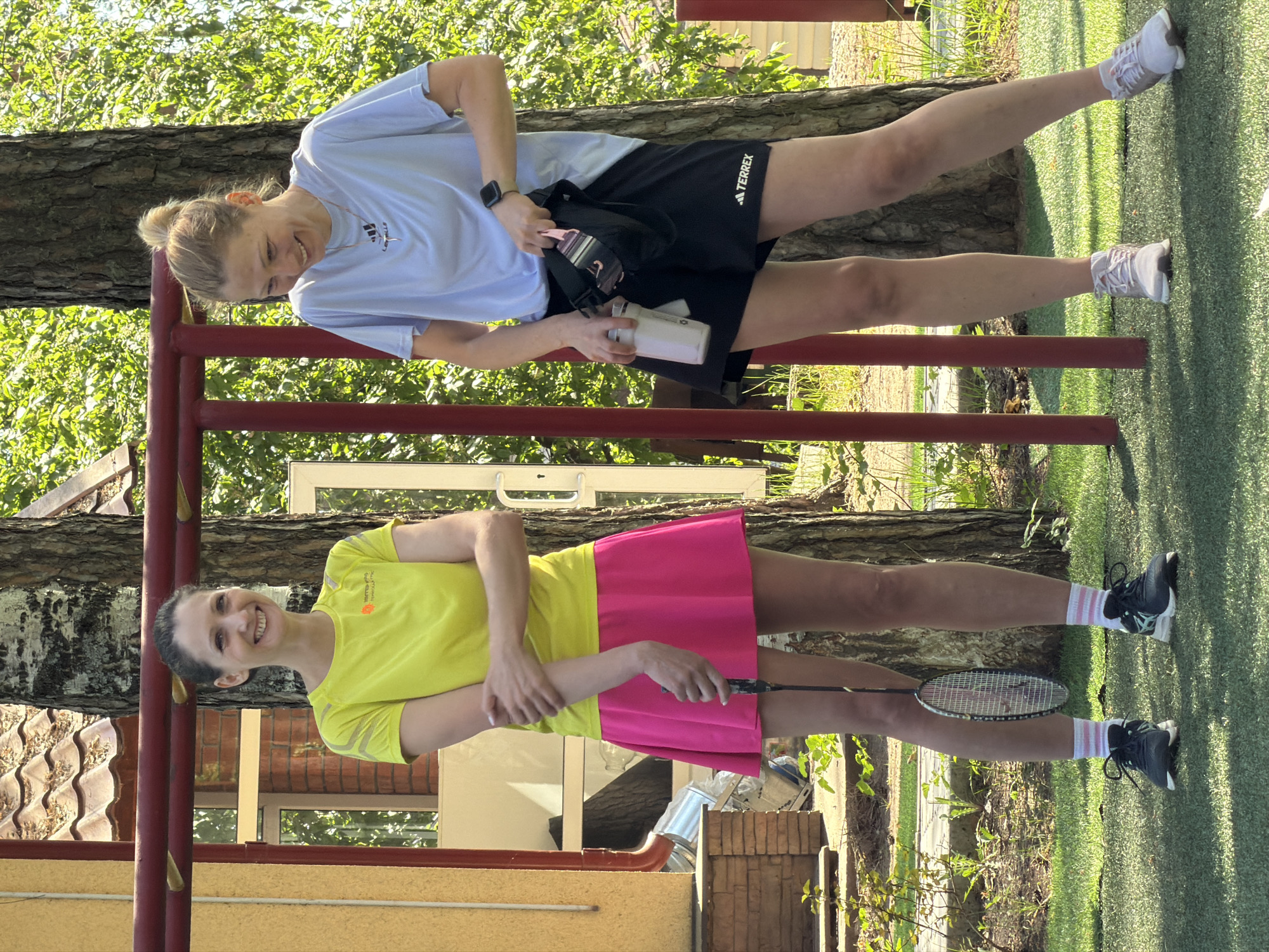 Two women stand outdoors by a red metal structure, possibly a climbing frame. The woman in the foreground wears a neon yellow shirt and a bright pink tennis skirt and holds a badminton racket. The woman in the background wears a light blue t-shirt, black shorts with "TERREX" printed on them, and white sneakers; she holds a water bottle and a black bag. The setting appears to be an outdoor recreation area or park, with grass and trees visible. A house or building is partially visible in the background. Based on the location clues, it is potentially in Chaykovsky, Russia.