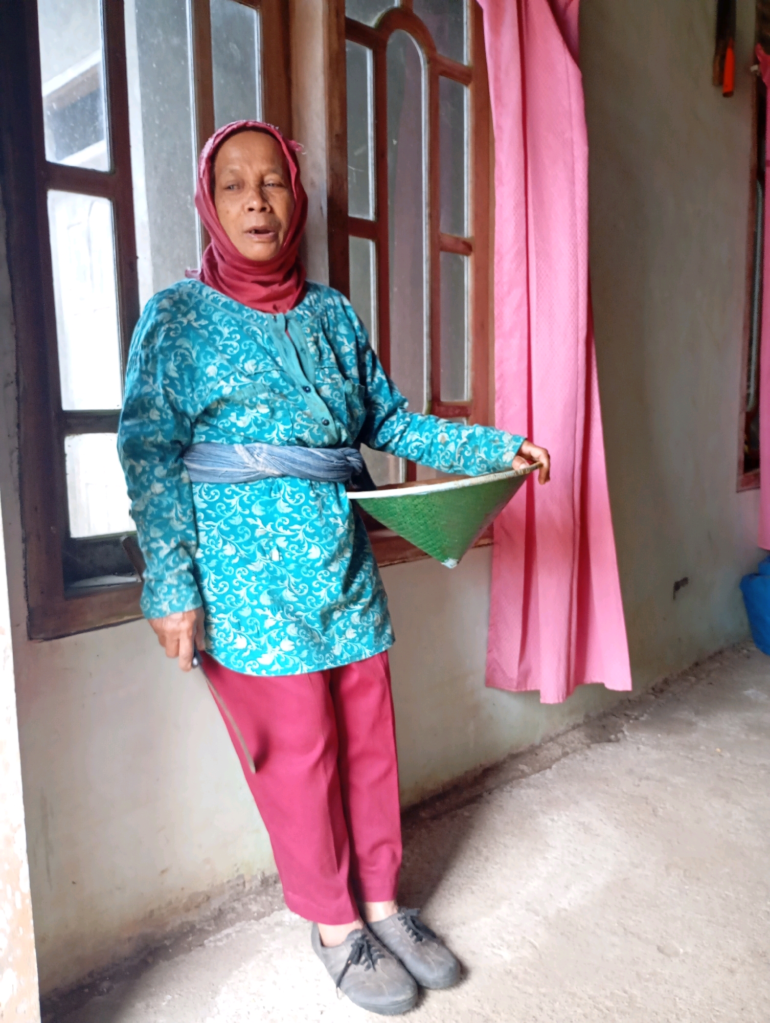 An elderly Indonesian woman, wearing a red headscarf and a teal patterned blouse with a grey sash, stands indoors. She is holding a conical green basket. The woman is positioned in front of a window with wooden frames and panes, partially obscured by pink curtains. The floor is made of concrete, and the wall is textured and light-colored. The scene suggests an interior residential setting in the city of Wonogondo, Indonesia.
