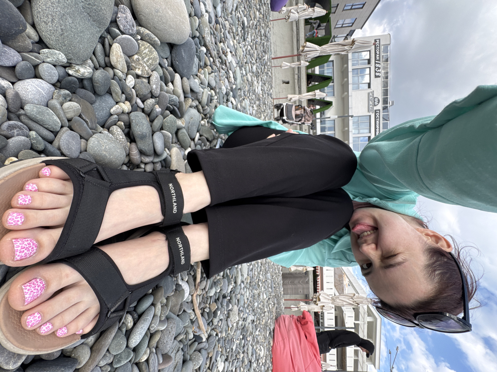 A woman is sitting on a pebble beach, wearing black pants, a light green hoodie, and black sandals. She is sticking out her tongue and winking at the camera. Her toes are painted with a pink leopard print design.
The background shows a building with a sign in Russian, identifying it as "RESTAURANT / 1838." Umbrellas and lounge chairs are visible near the building.
The location is likely Sochi, Russia, based on the Russian signage and beach environment.